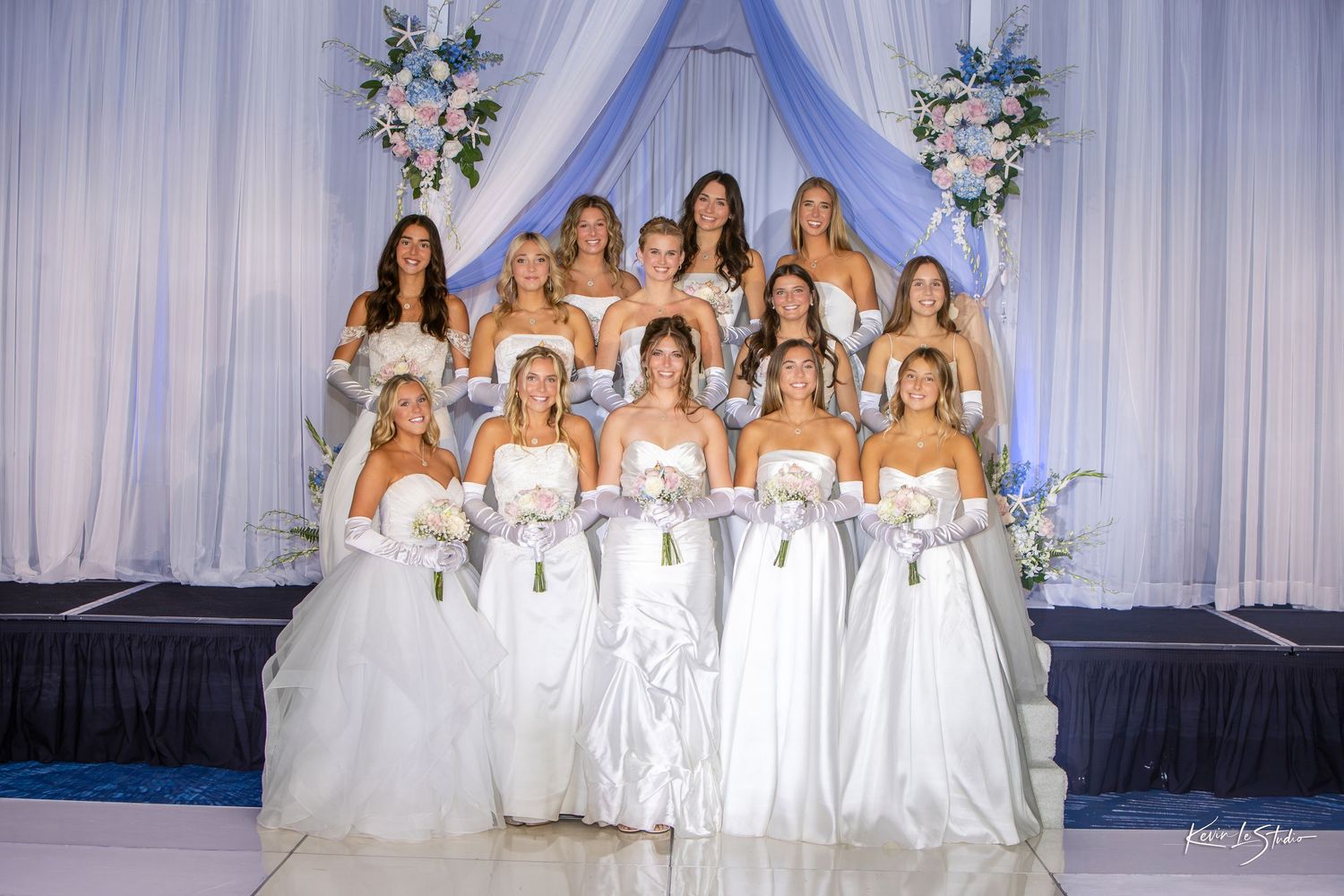 Group of elegant bridesmaids in white gowns and gloves posing on a decorated stage with blue and white floral backdrop.
