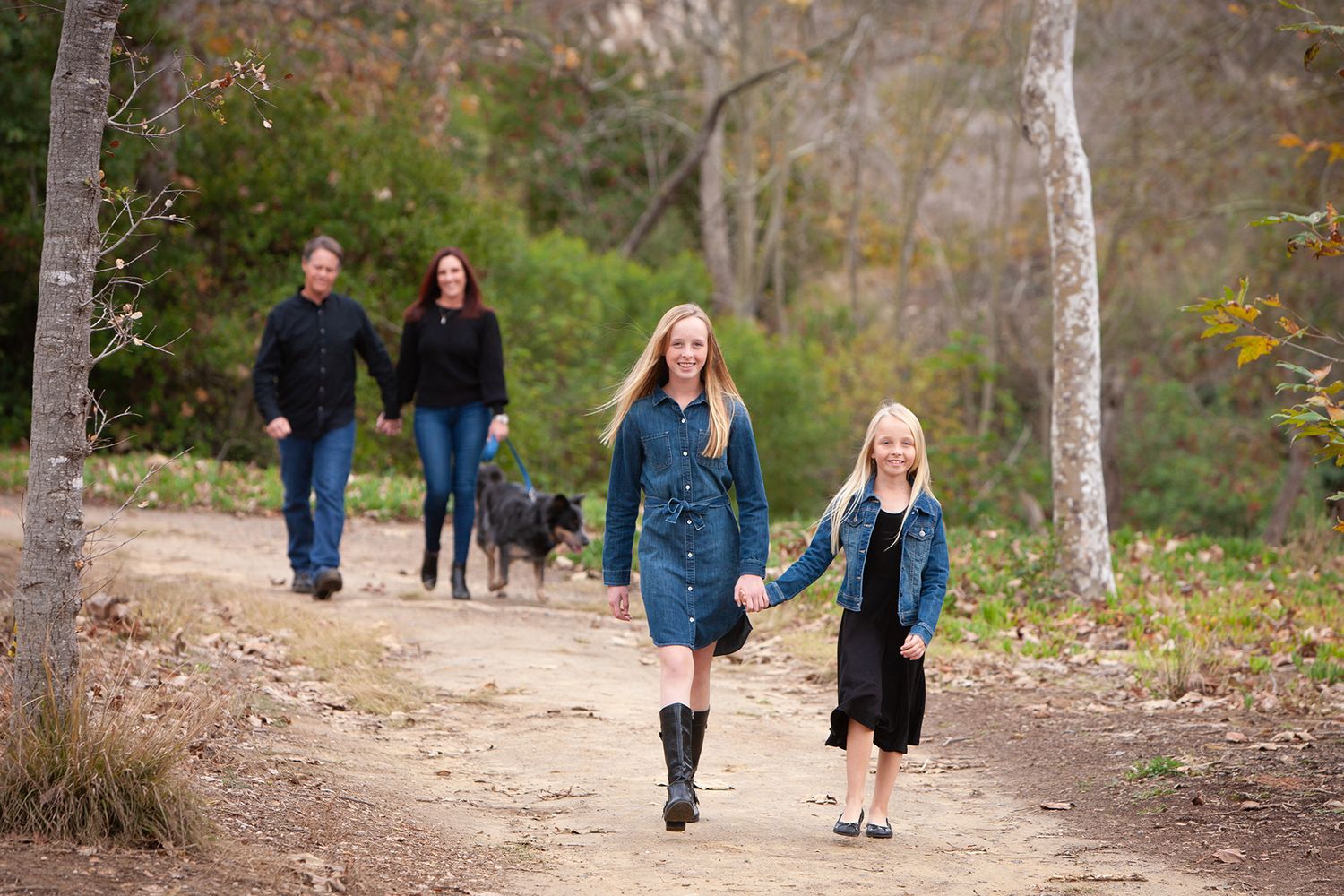 Family with two girls and a dog walking on a wooded trail in autumn, enjoying a casual outdoor stroll.