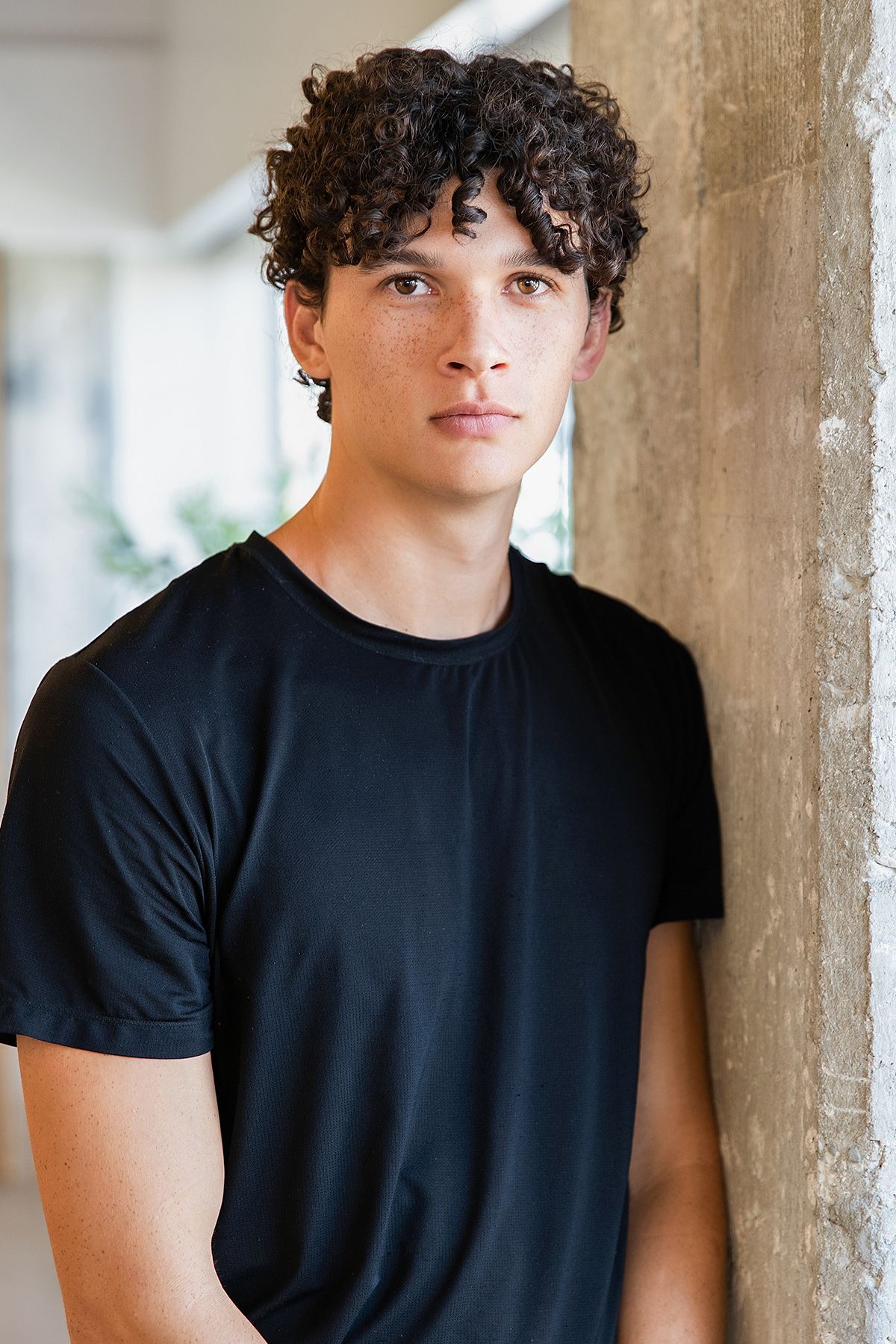 Young man with dark curly hair wearing a black t-shirt, leaning against a concrete wall indoors.