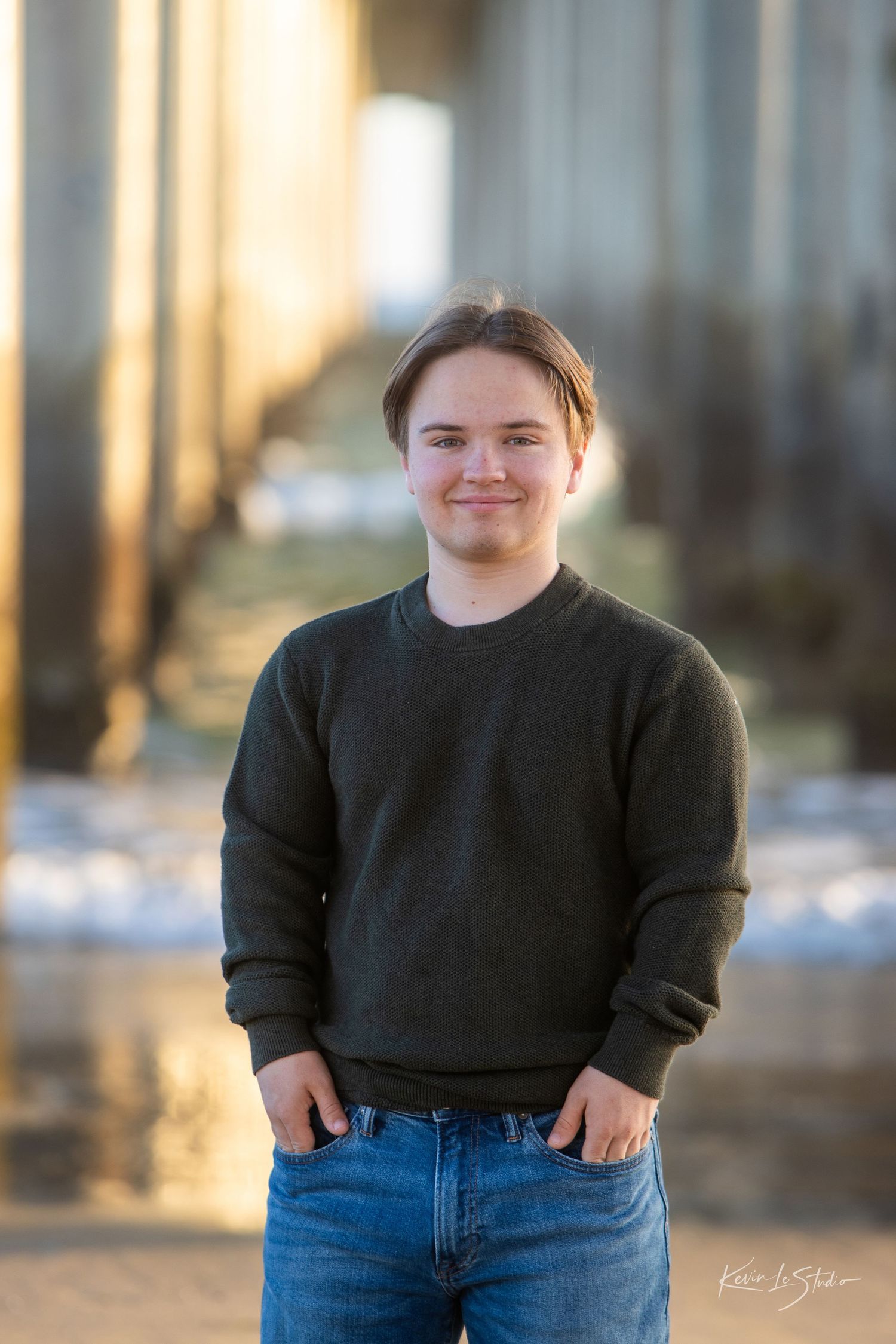Smiling young man in dark green sweatshirt and jeans posing outdoors.