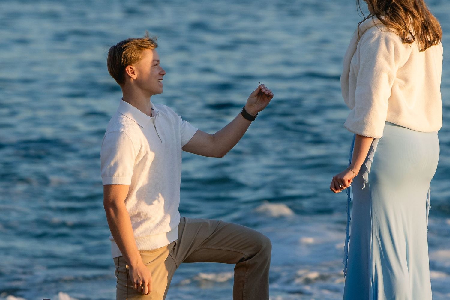 Young man on one knee proposing to woman by the ocean at golden hour.