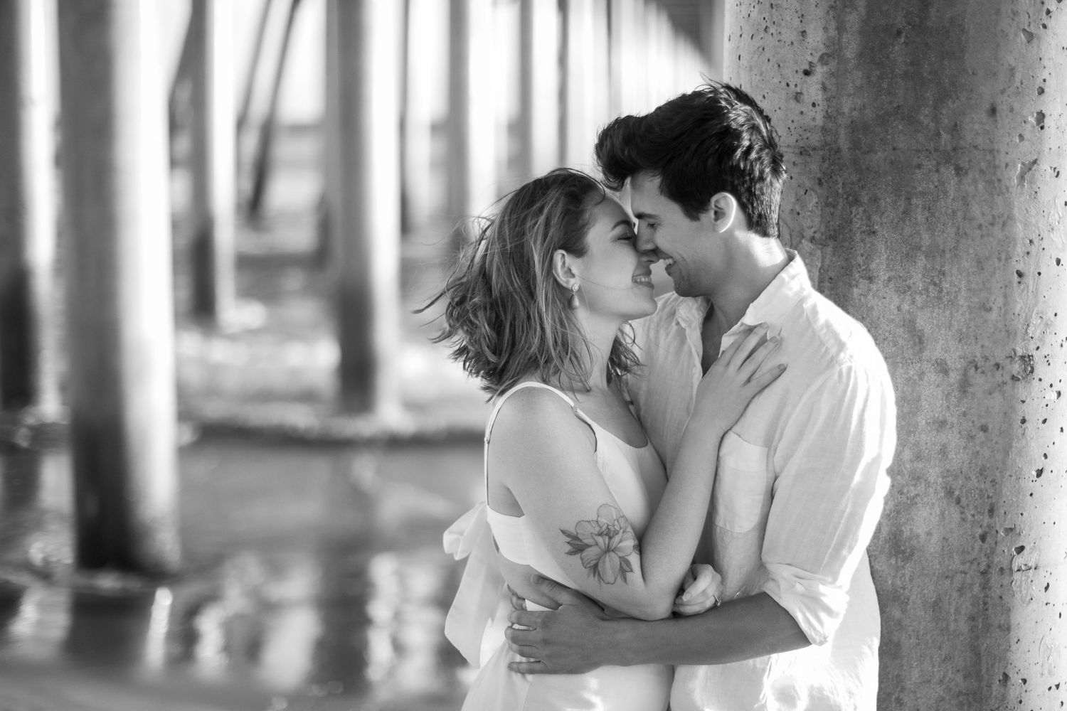 Black and white photo of a romantic couple almost kissing under a pier near the ocean.