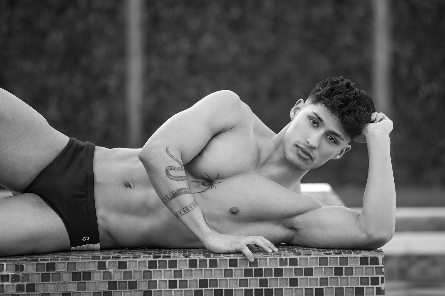 Black and white photo of tattooed male model in swim briefs by a tiled pool.