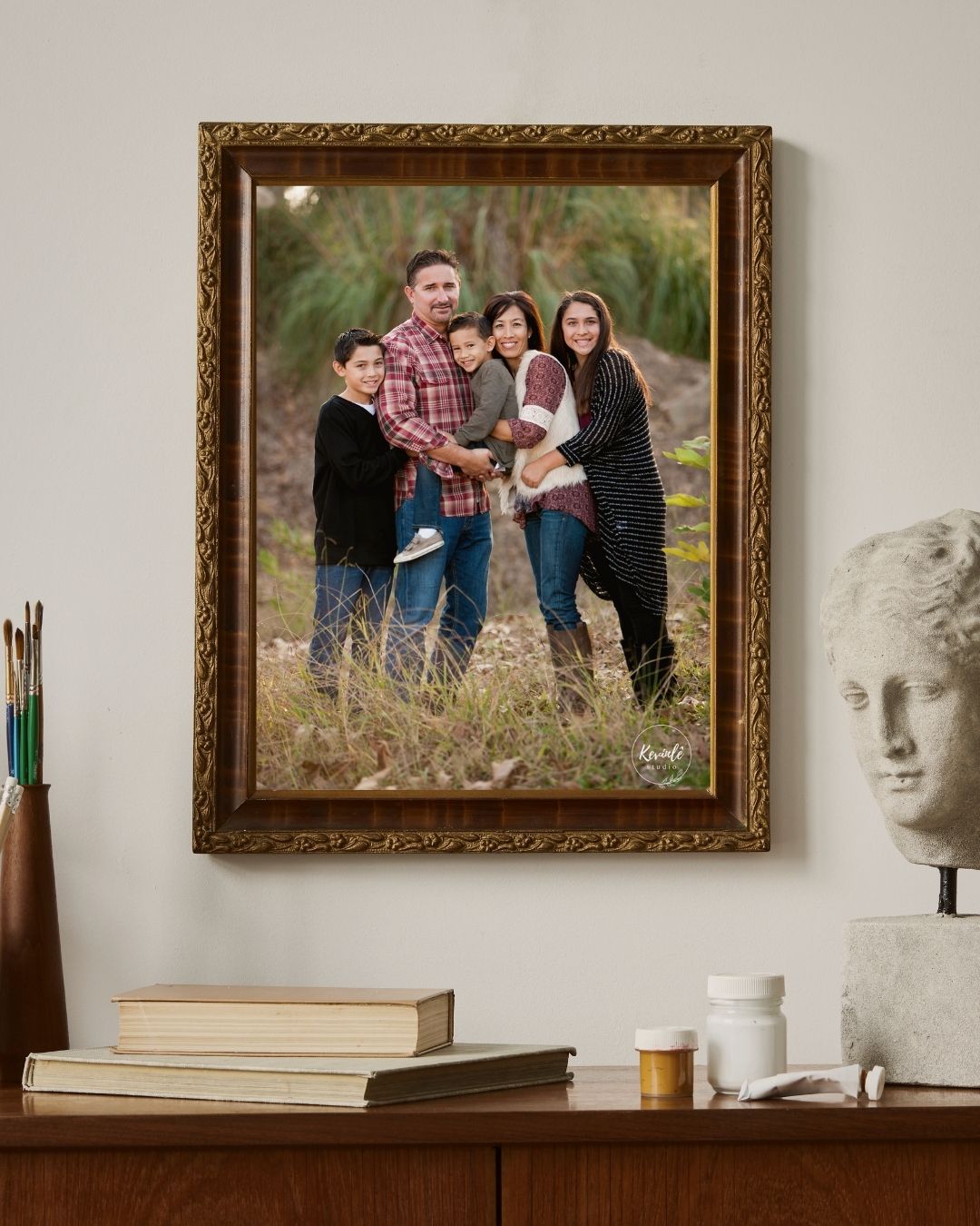 Ornate gold-framed family portrait of five people posing outdoors in a field, displayed on a wall above a wooden dresser.