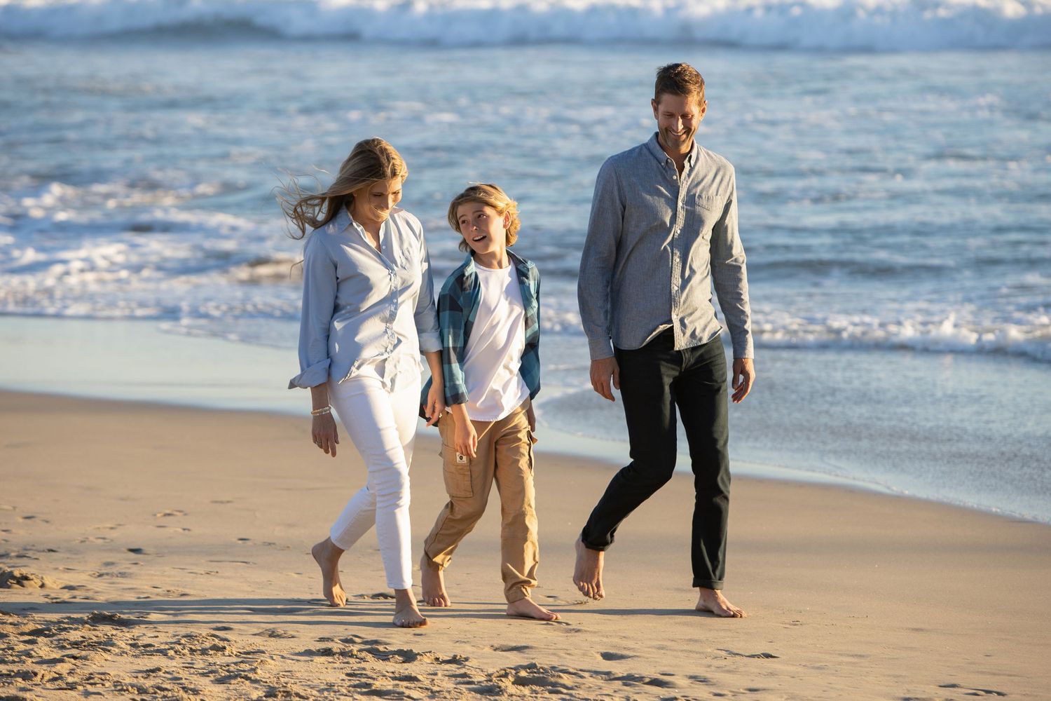 Happy family of three walking barefoot on a sunny beach, with ocean waves in the background.