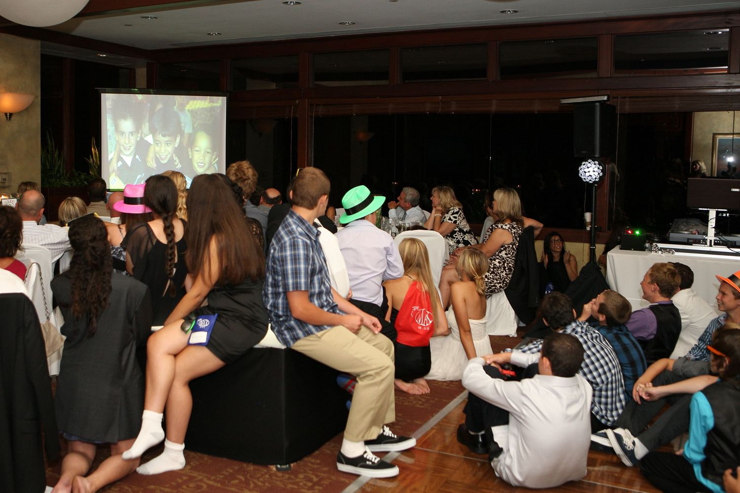 Guests gather at an indoor party venue watching a projected screen, wearing colorful hats and festive attire.