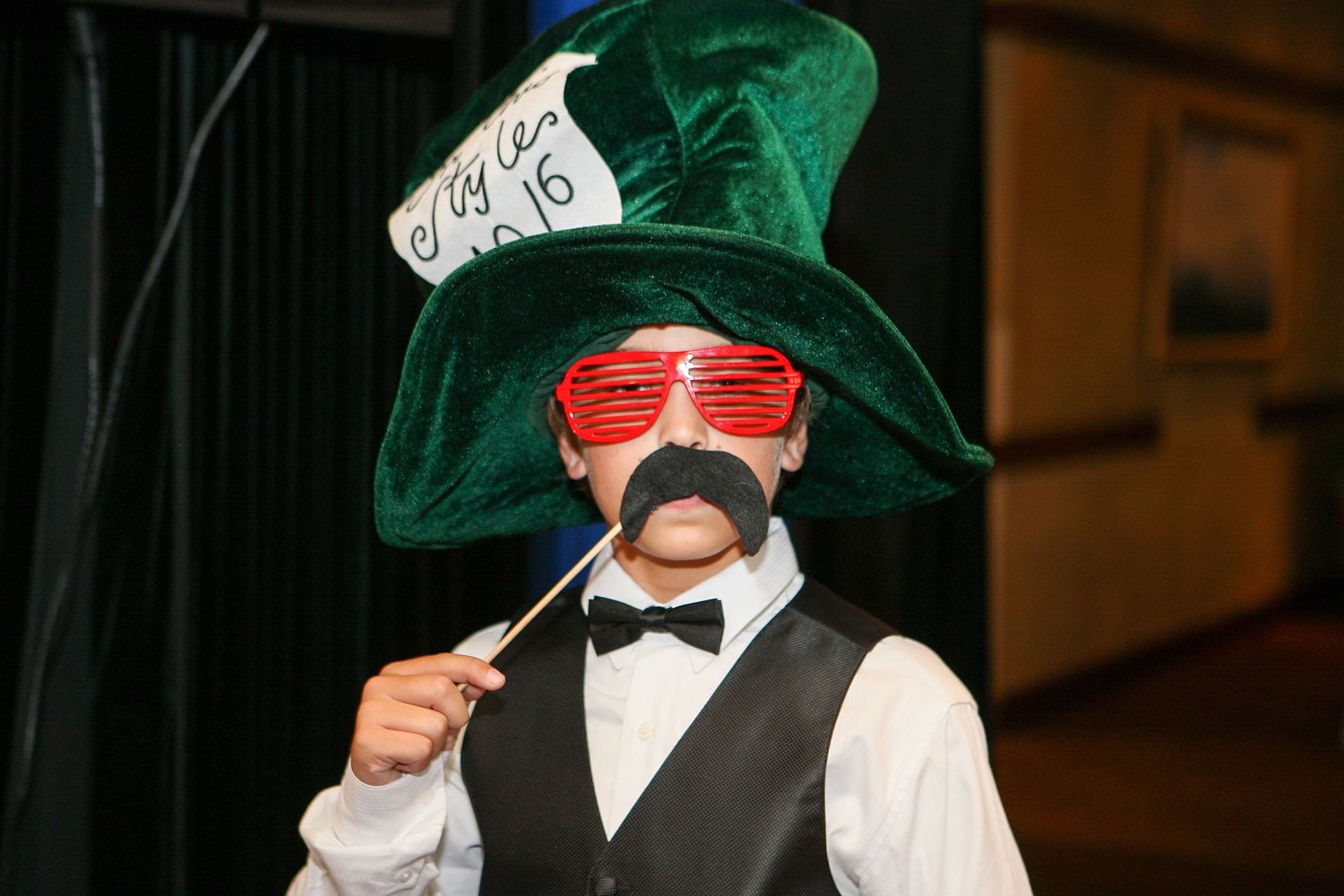 Person wearing green Mad Hatter top hat, red oversized glasses, and fake mustache at a costume event.