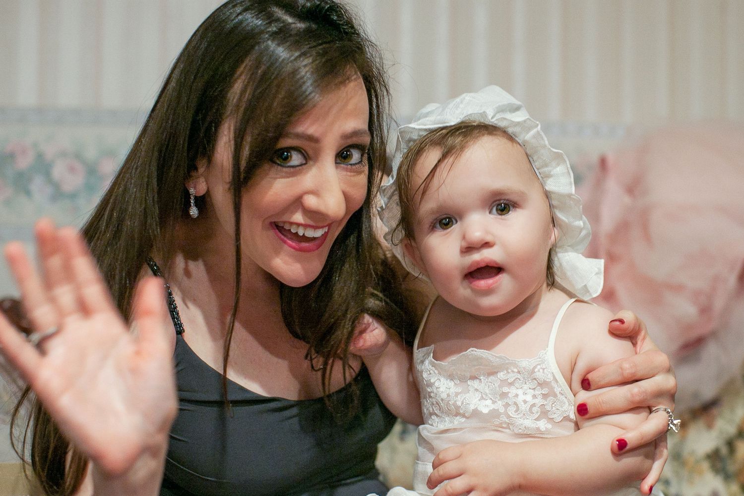 A smiling woman with dark hair holds a baby girl in a white dress and bonnet, waving at the camera indoors.