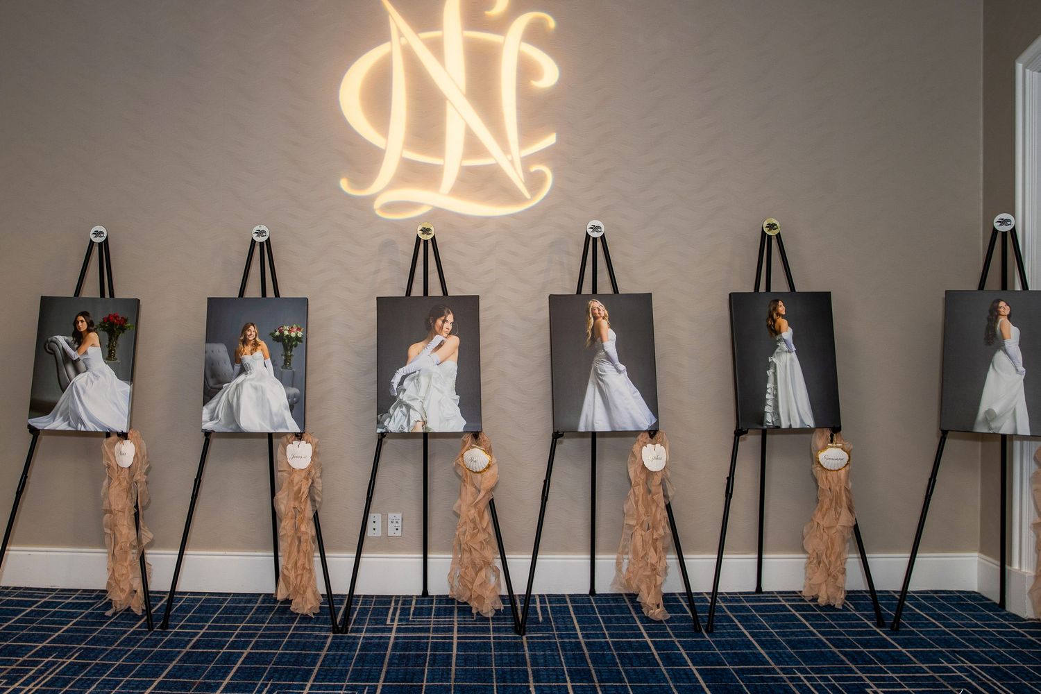 Six bridal portrait photos displayed on black easels with ribbon bows beneath a glowing monogram sign at a wedding venue.