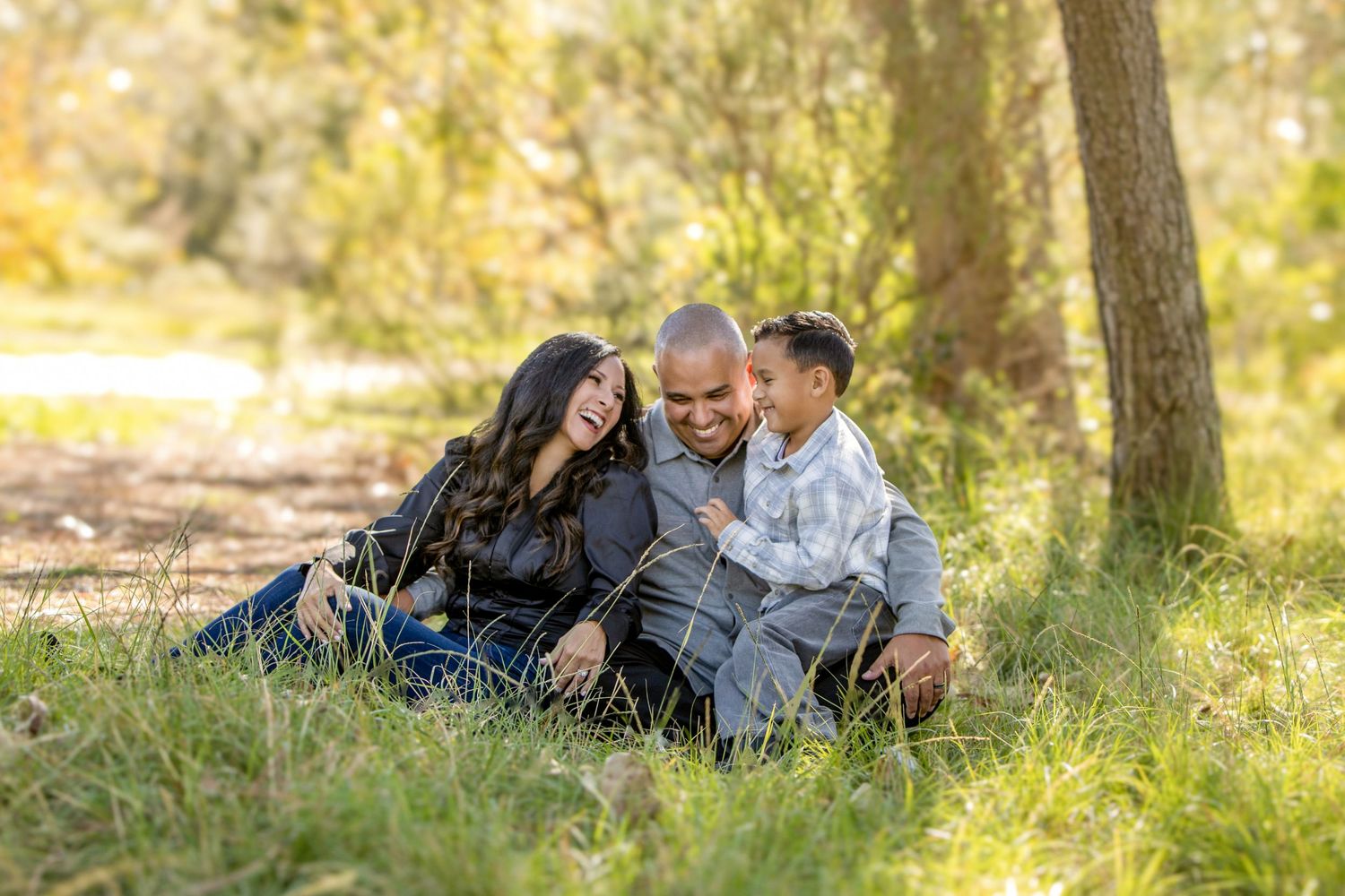 Happy family of three sitting together in lush green grass, laughing and bonding outdoors near trees.