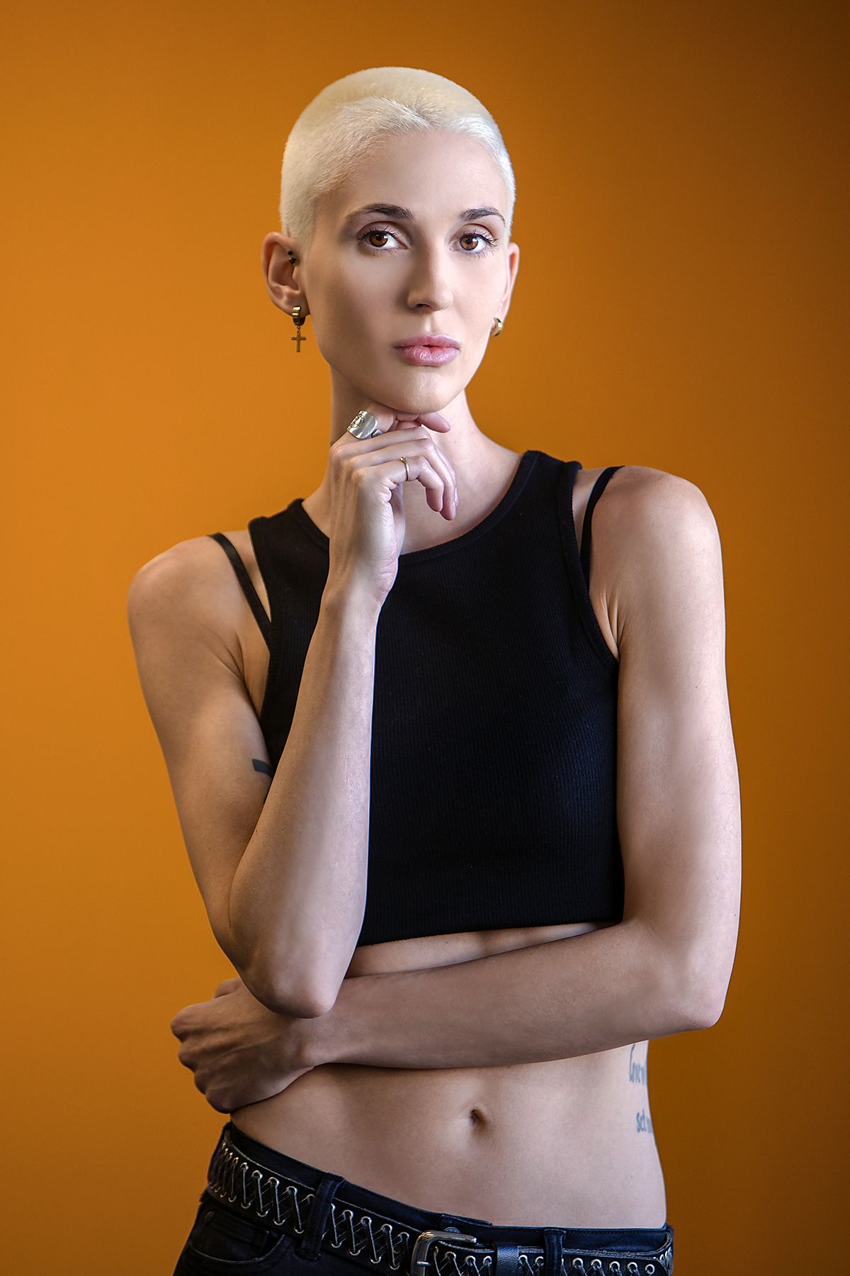 Edgy woman with buzzed blonde hair in black crop top poses thoughtfully against orange background.