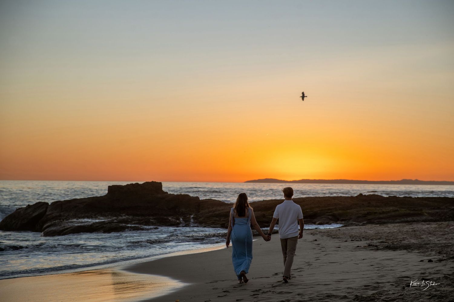 Couple holding hands walking along beach at sunset with bird flying overhead.