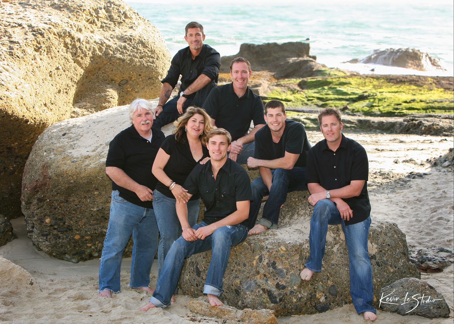 Family of seven posing on beach rocks in matching black shirts for a professional coastal photo session.