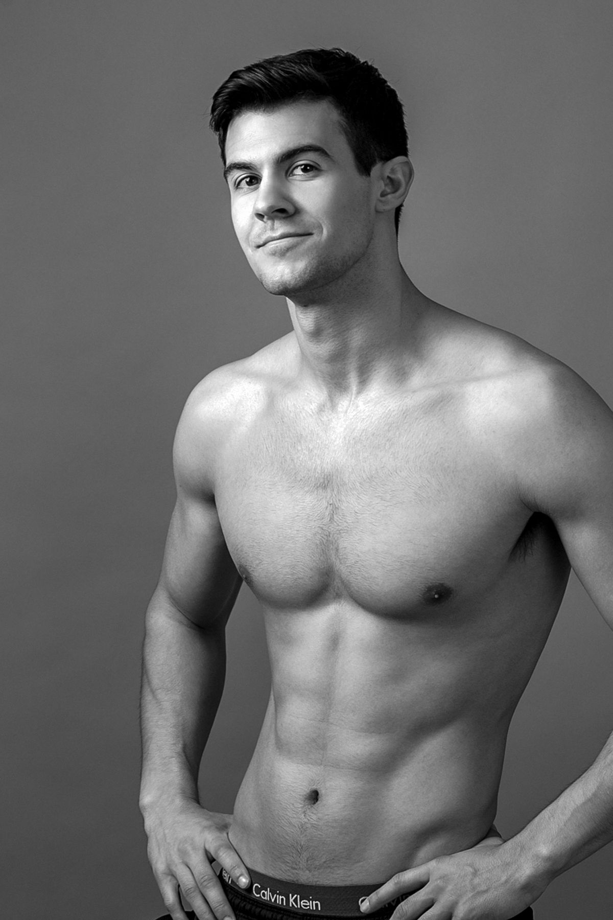 Black and white photo of a fit shirtless man wearing Calvin Klein underwear posing confidently against a gray background.