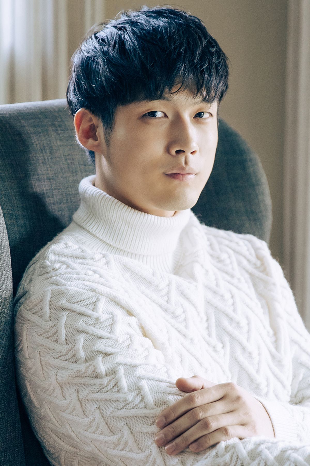 Young Asian man in a white cable-knit turtleneck sweater sitting in a gray armchair.