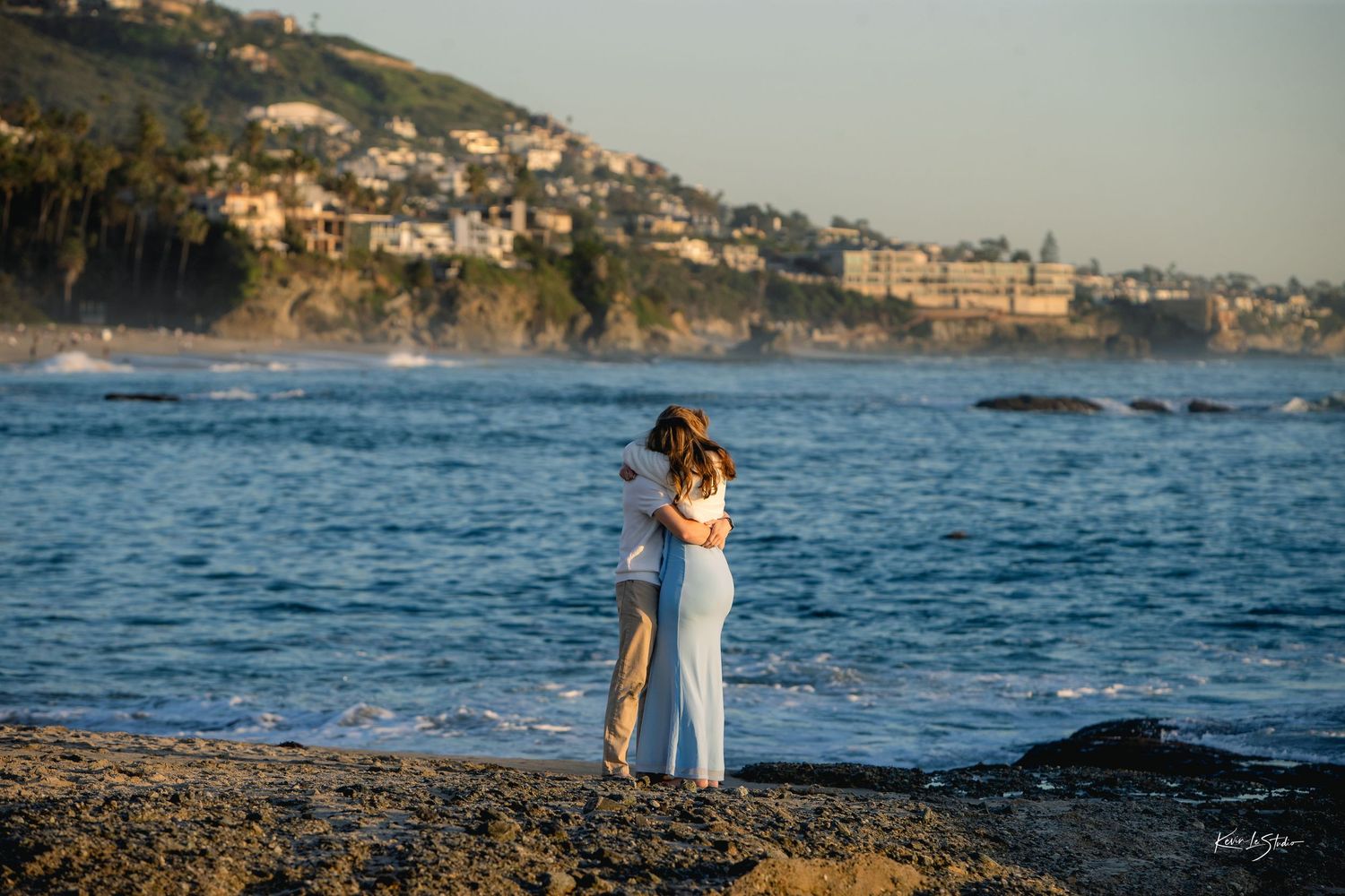 Couple embracing on rocky beach with scenic coastal hillside in background.