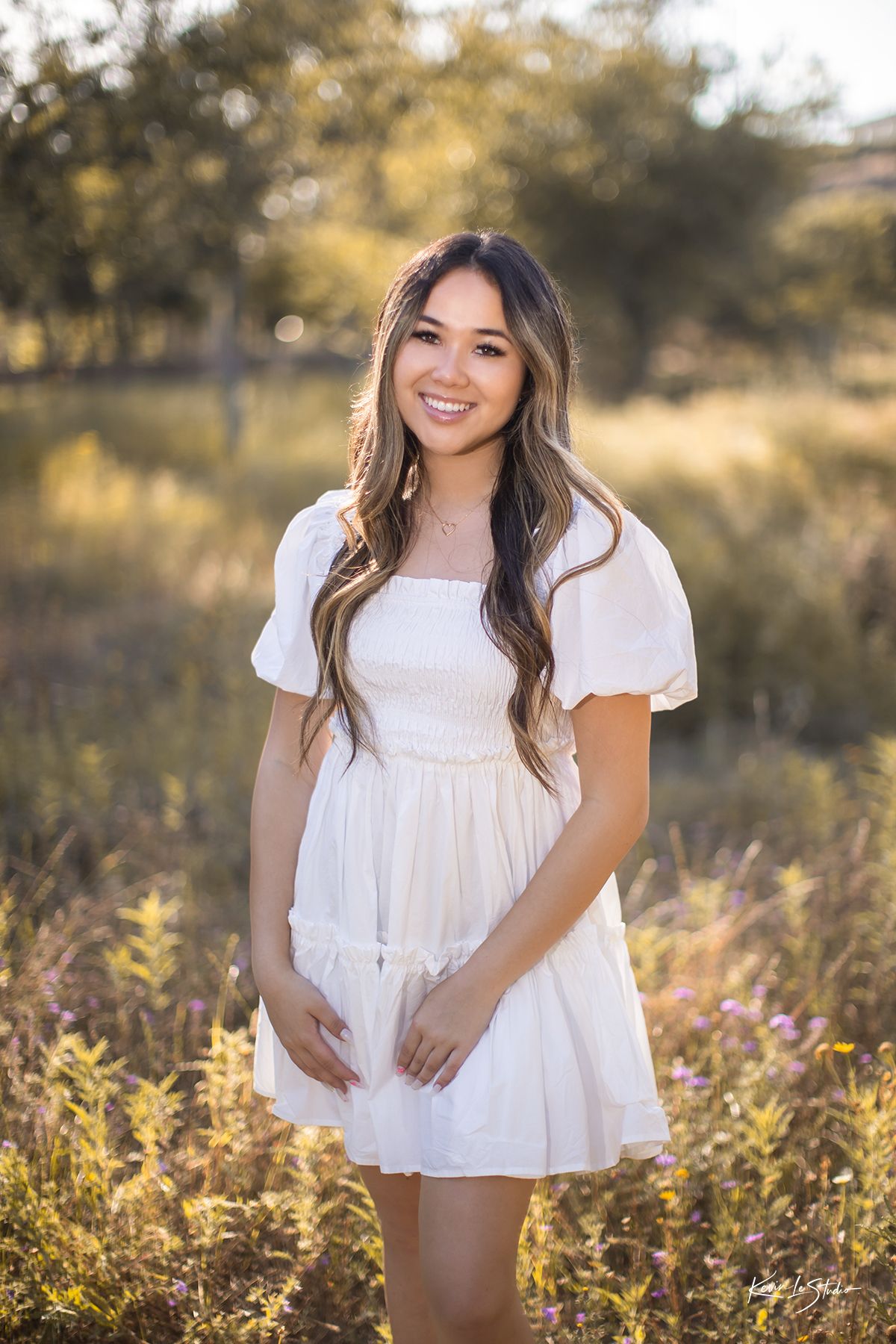 Smiling young woman in white mini dress stands in sunlit meadow during outdoor senior portrait photography session.
