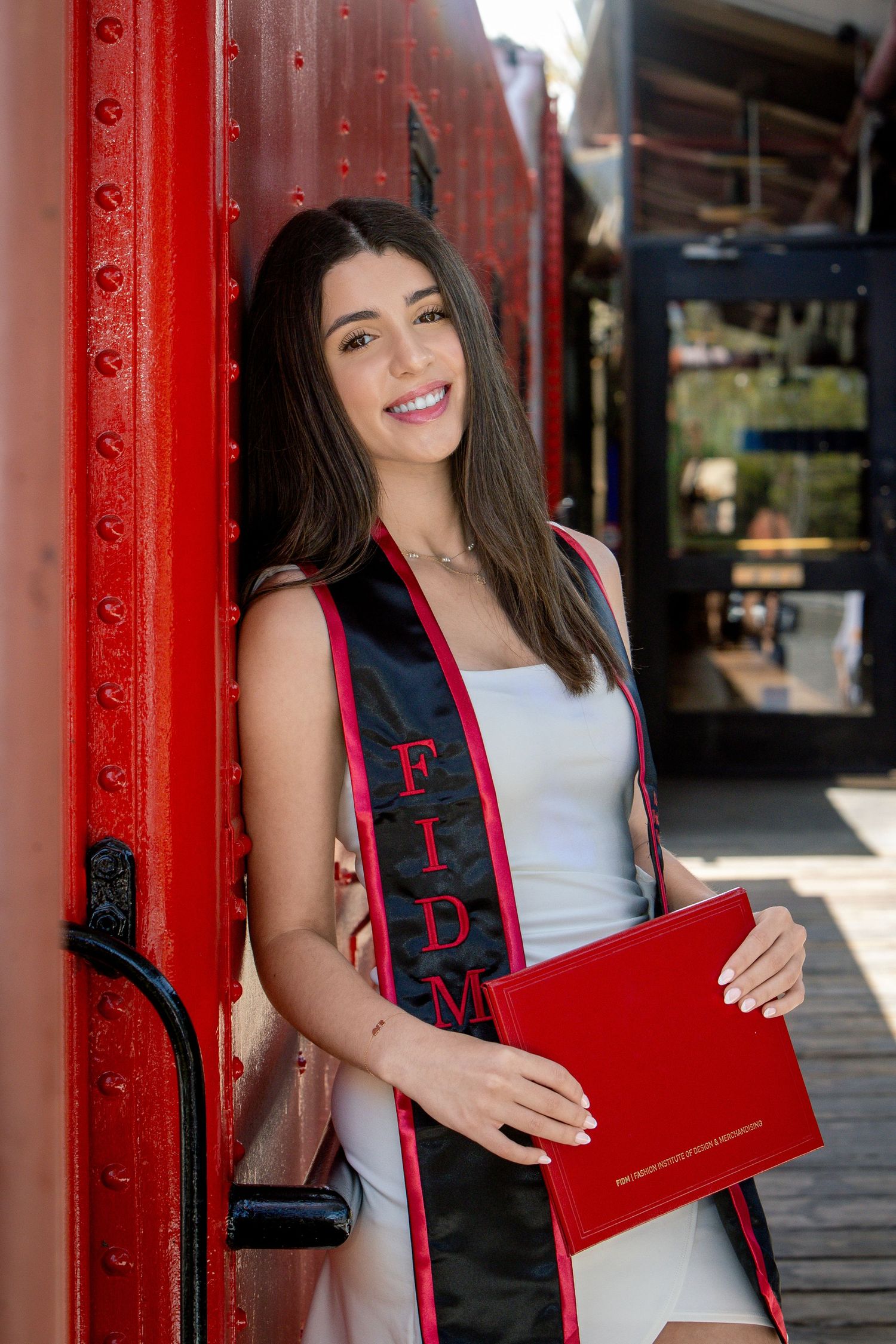 Female FIDM graduate smiles holding red diploma while leaning against vibrant red train car in white dress.
