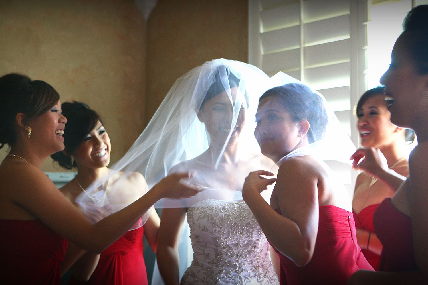 Bridesmaids in red dresses helping bride with white veil and lace wedding gown indoors.