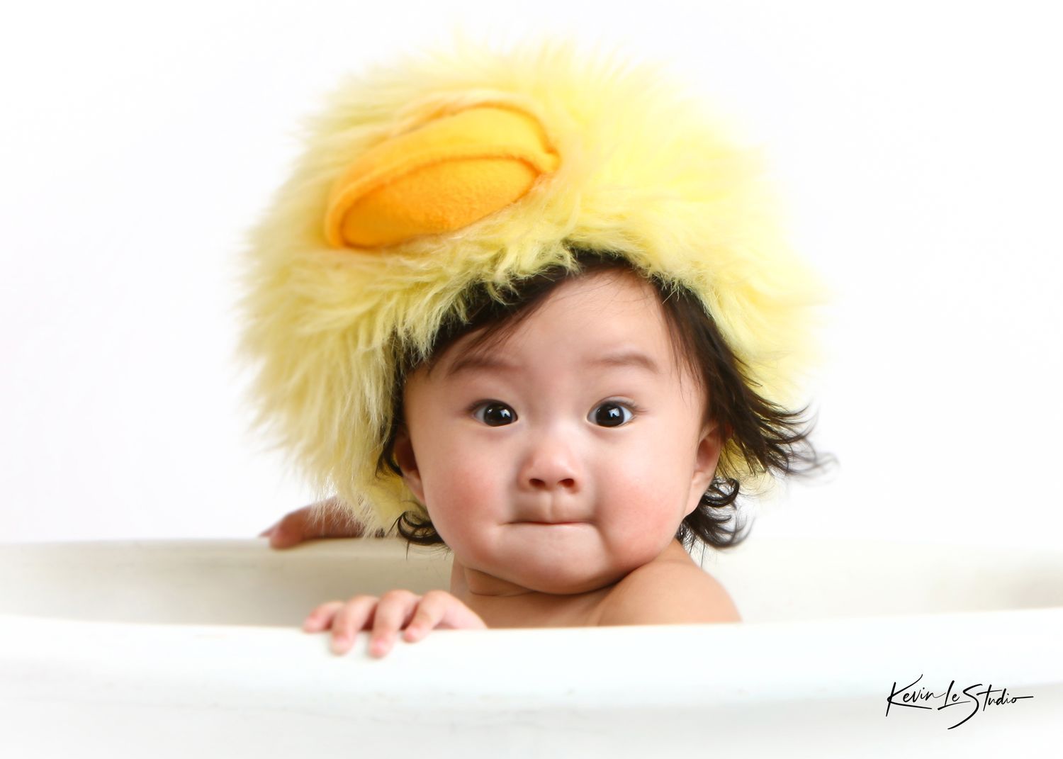 A cute baby wearing a fluffy yellow duck hat peeks over the edge of a white bathtub.