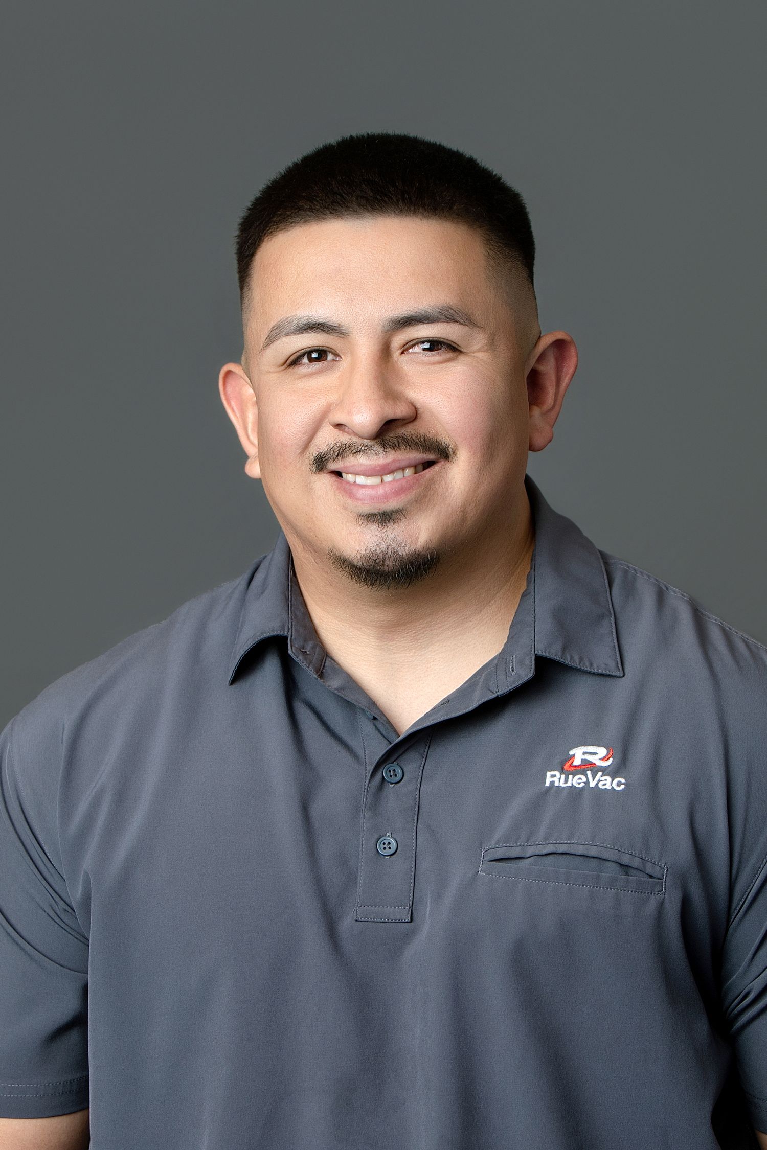 Professional headshot of a smiling man wearing a gray RueVac polo shirt against a gray background.