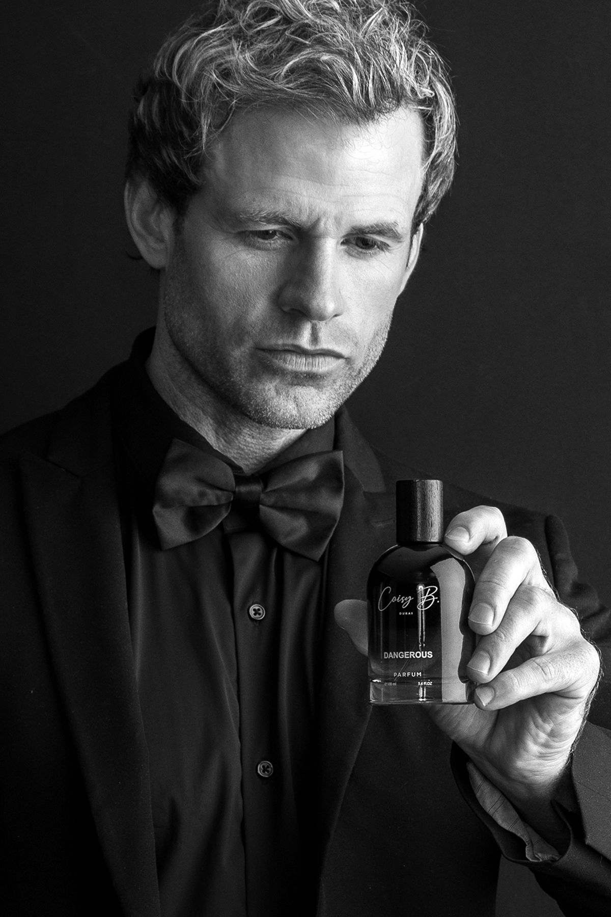 Black and white photo of a man in a bow tie holding a cologne bottle labeled Dangerous.