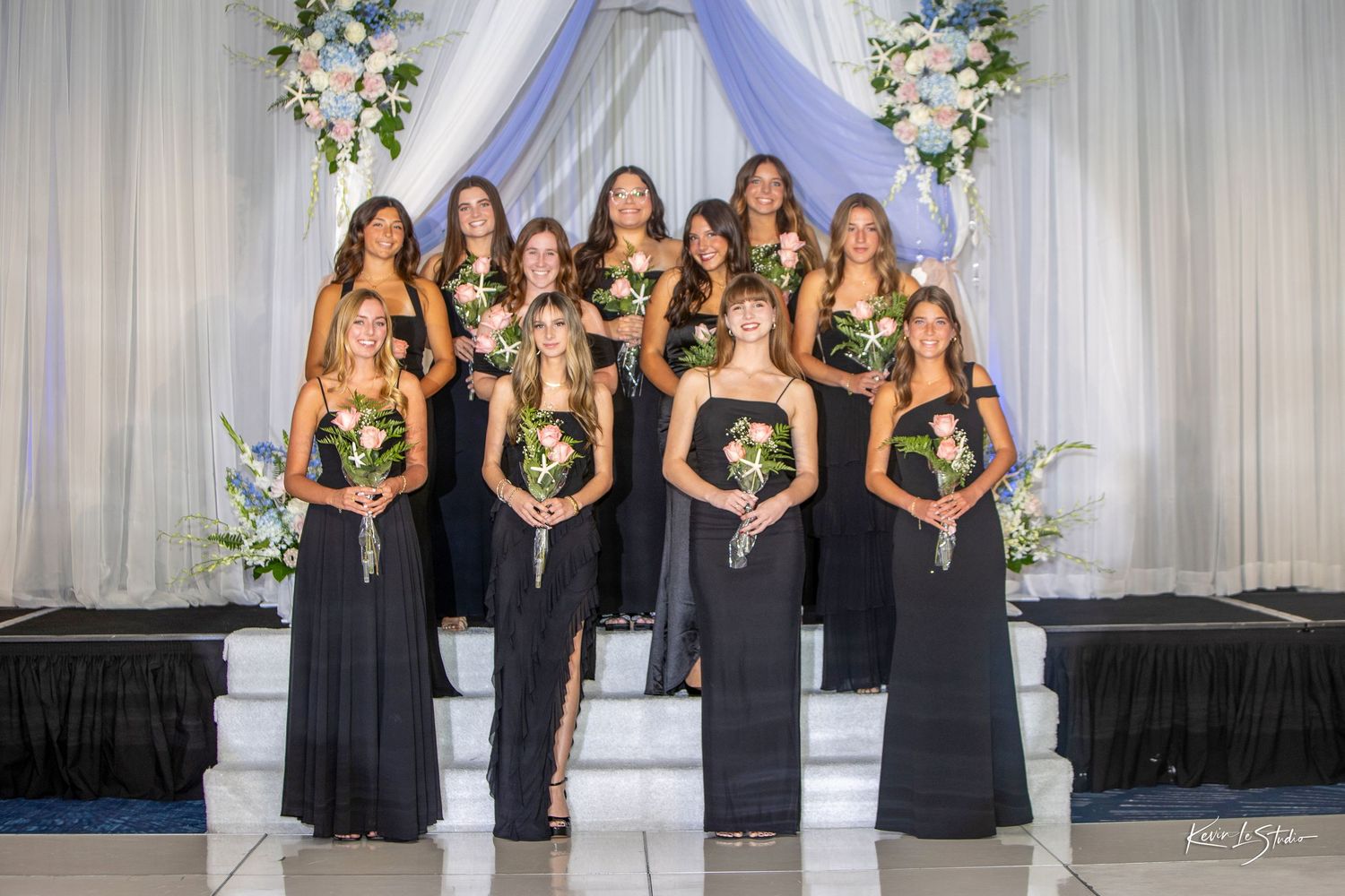 Nine bridesmaids in black gowns holding bouquets pose on steps before a floral wedding altar backdrop.