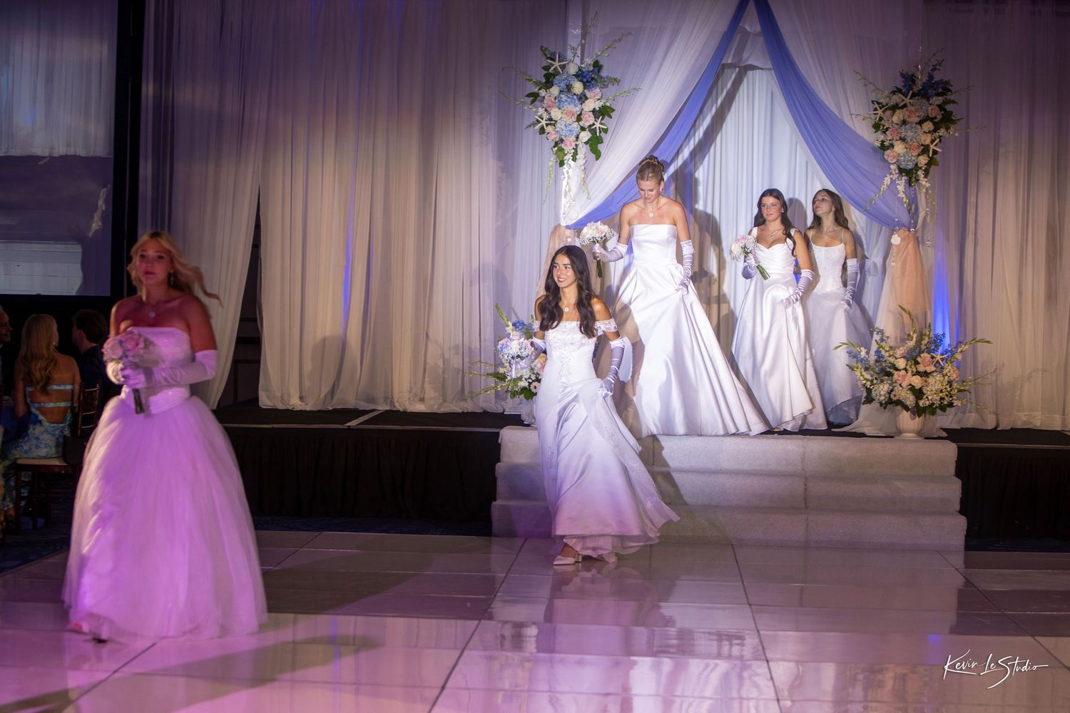 Elegant bridal fashion show with models in white wedding gowns walking a lit stage with floral arch backdrop.