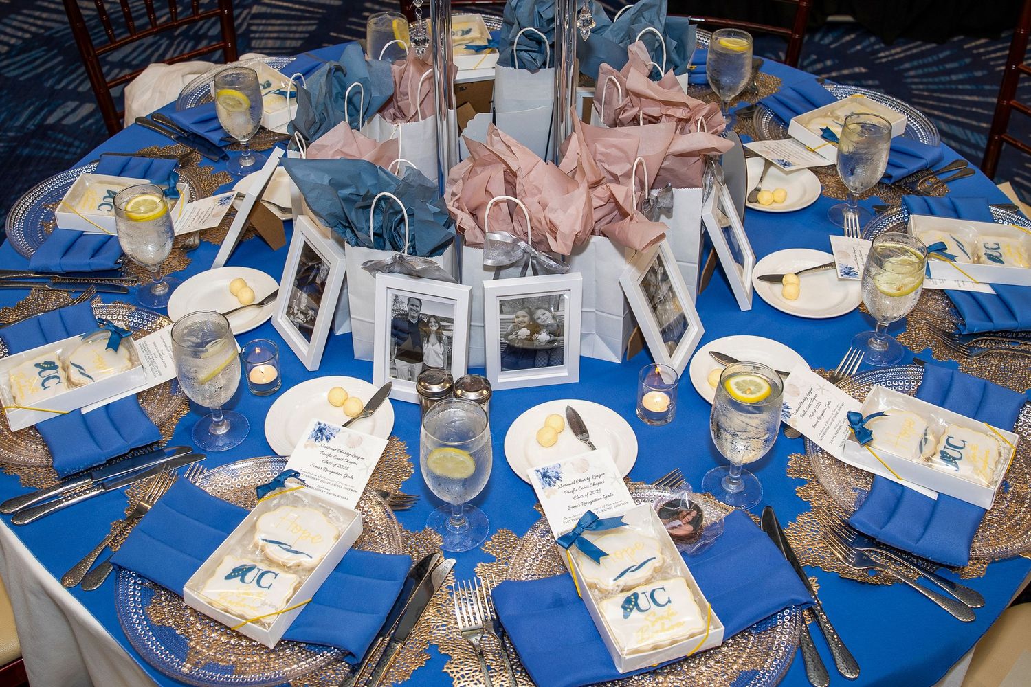 Elegant banquet table set with blue napkins, gold chargers, framed photos, and gift bags for a formal event.