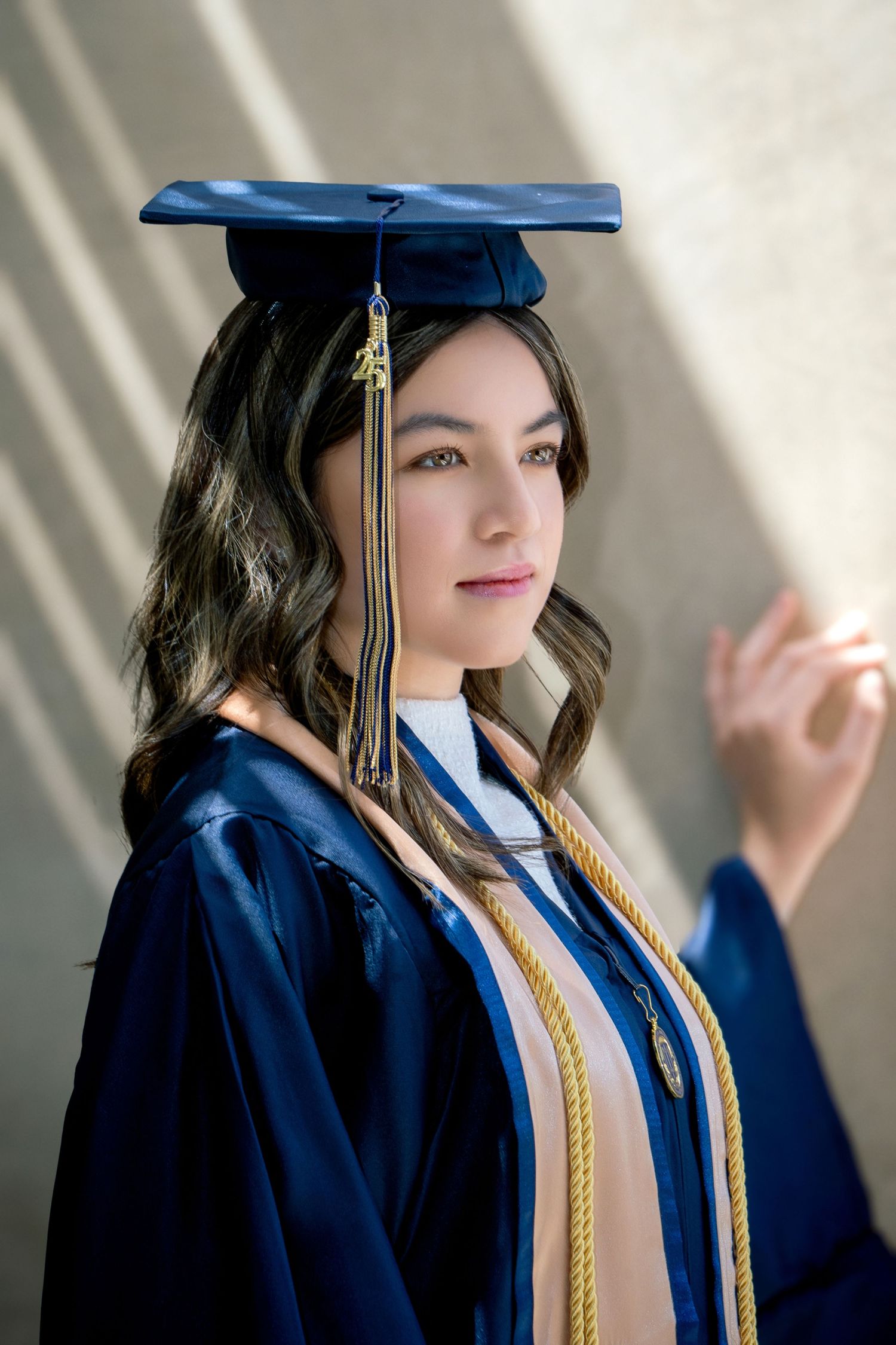 Young woman in navy blue graduation cap and gown with gold honor cords poses proudly for graduation portrait.