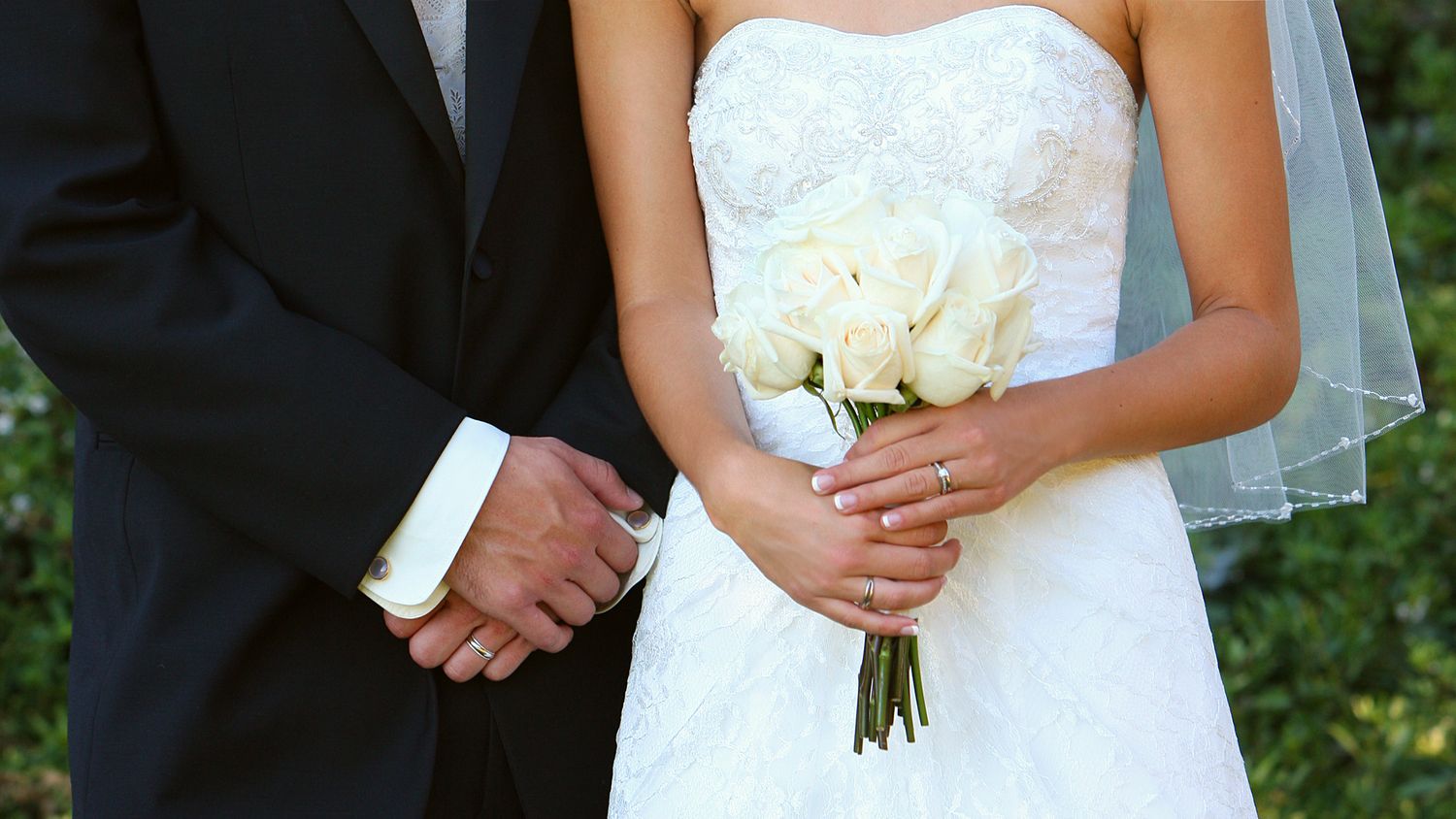 Bride and groom holding hands, bride wearing white lace dress and veil, holding white rose bouquet.