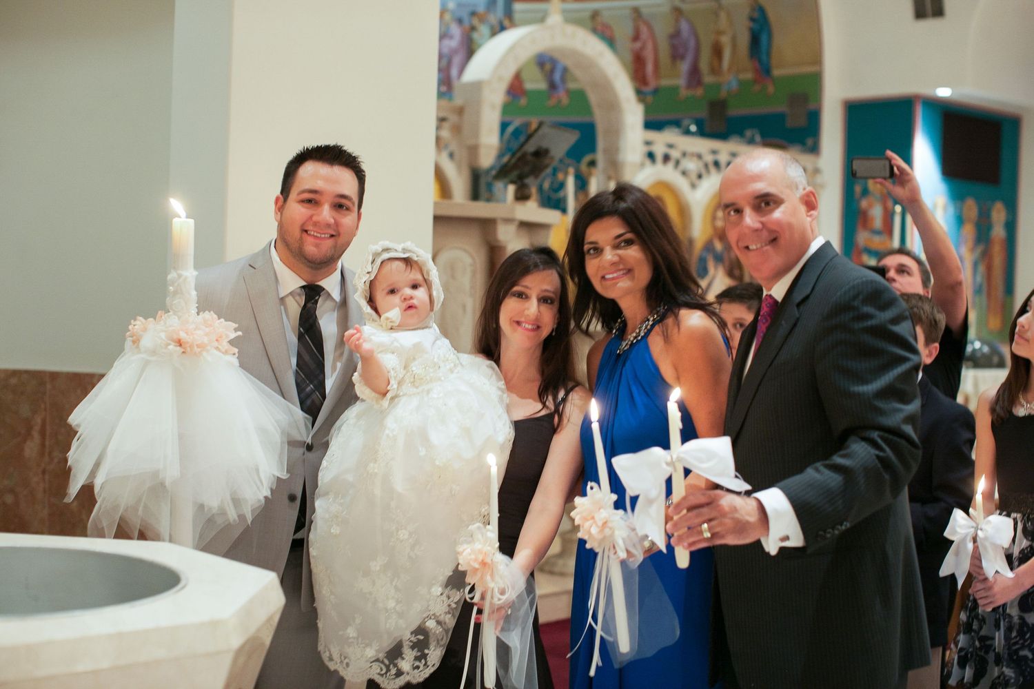 Family poses with baby in christening gown during baptism ceremony in church, holding decorated candles.