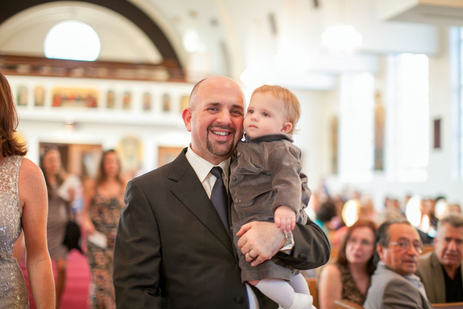 Smiling bald man in suit holding a baby in gray outfit inside a church during a wedding ceremony.