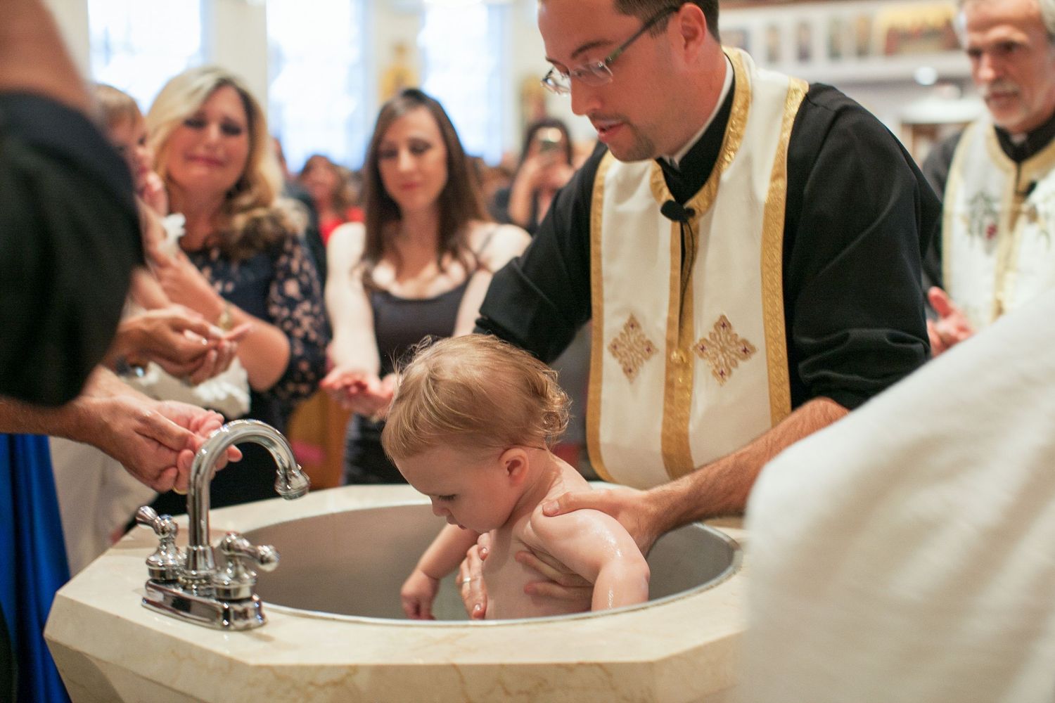Priest performing infant baptism ceremony at a church font, with family members gathered around watching.