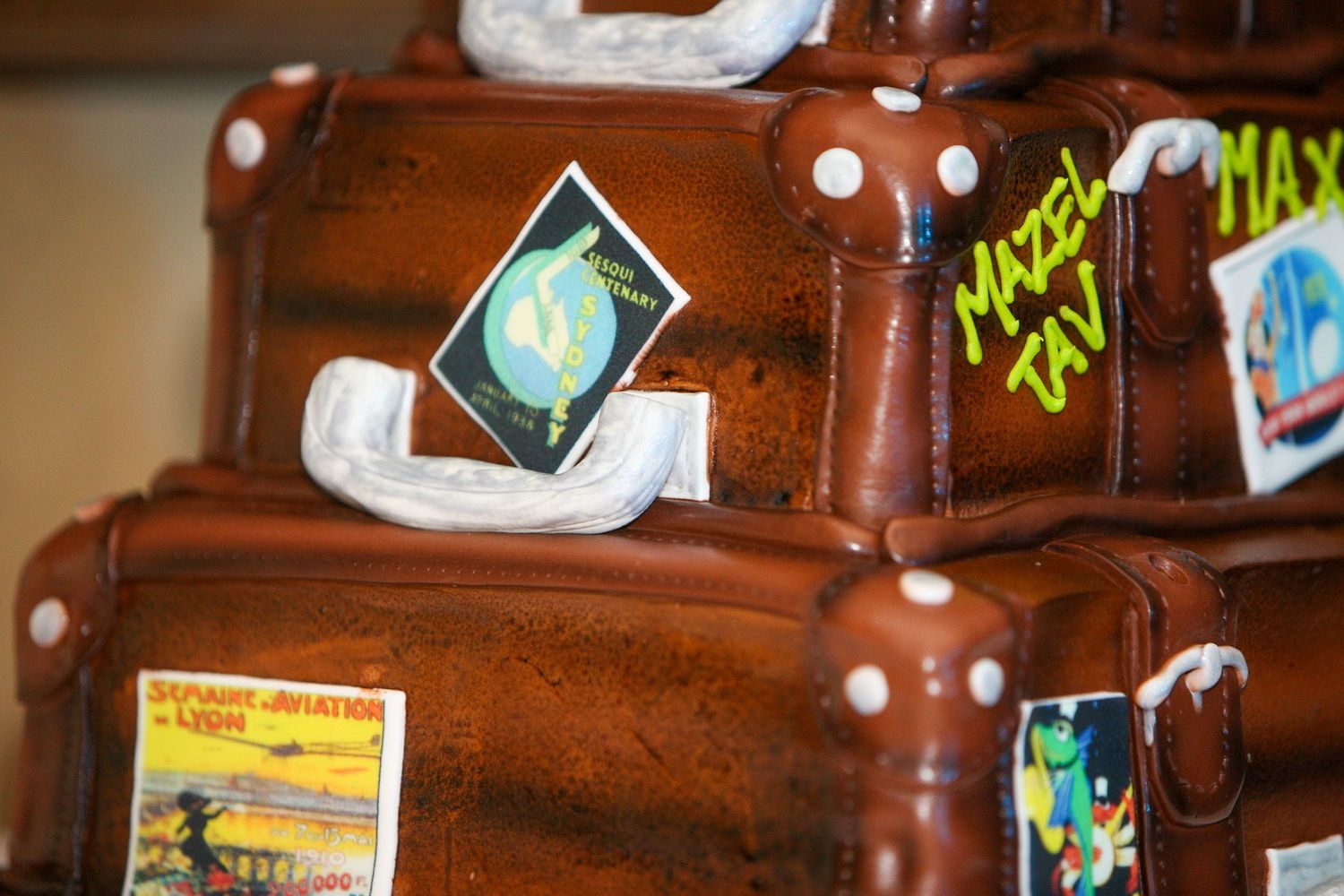 Stacked suitcase-shaped chocolate cake decorated with travel stickers and Mazel Tov text for a celebration.