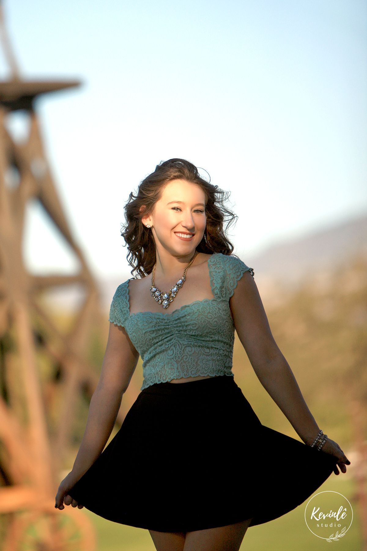 Young woman in teal top and black skirt smiling outdoors, wearing a statement necklace in a sunny field.