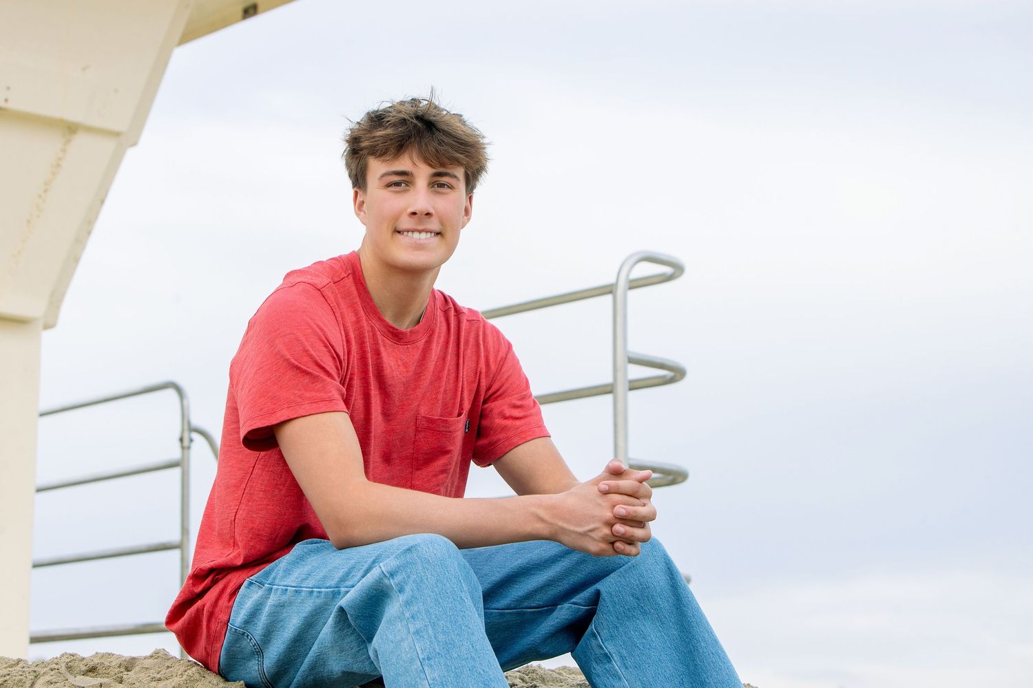 Smiling teenage boy in red t-shirt and jeans sitting outdoors near metal railing against cloudy sky.