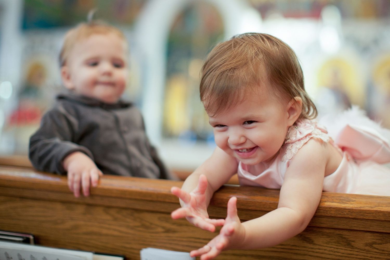 Two adorable babies in a church pew, girl in pink tutu smiling and reaching forward at a baptism.