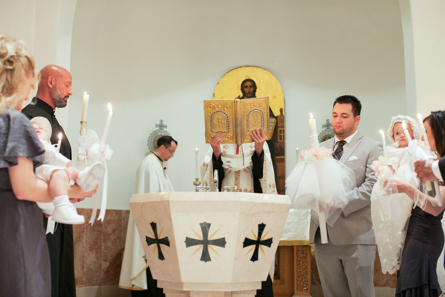 Priest performs a baptism ceremony at a church font, with godparents holding infants in white gowns.