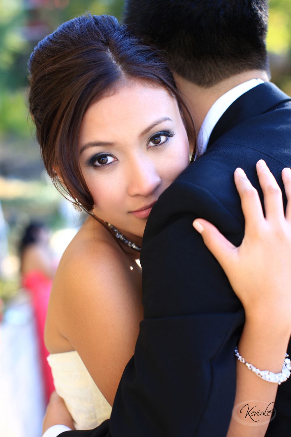 A bride in a strapless gown embraces a man in a dark suit, gazing over his shoulder in an outdoor wedding portrait.