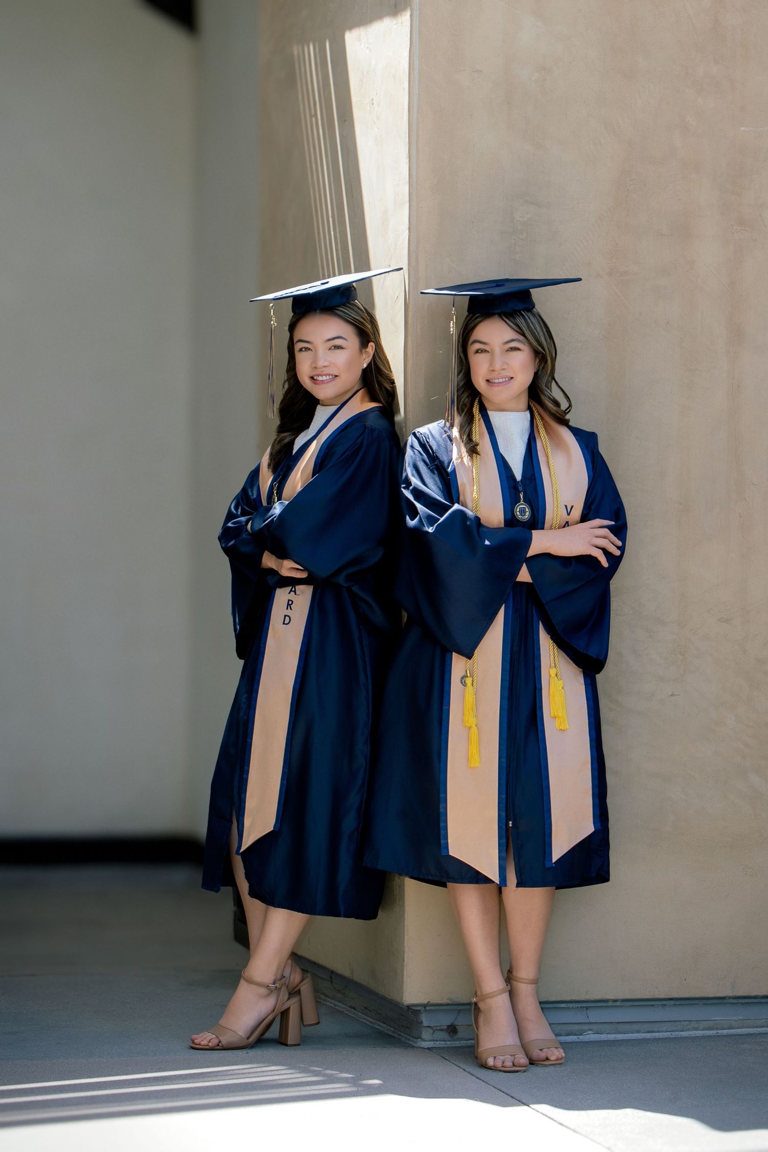 Two smiling graduates in navy blue cap and gown pose back-to-back with arms crossed at their graduation ceremony.