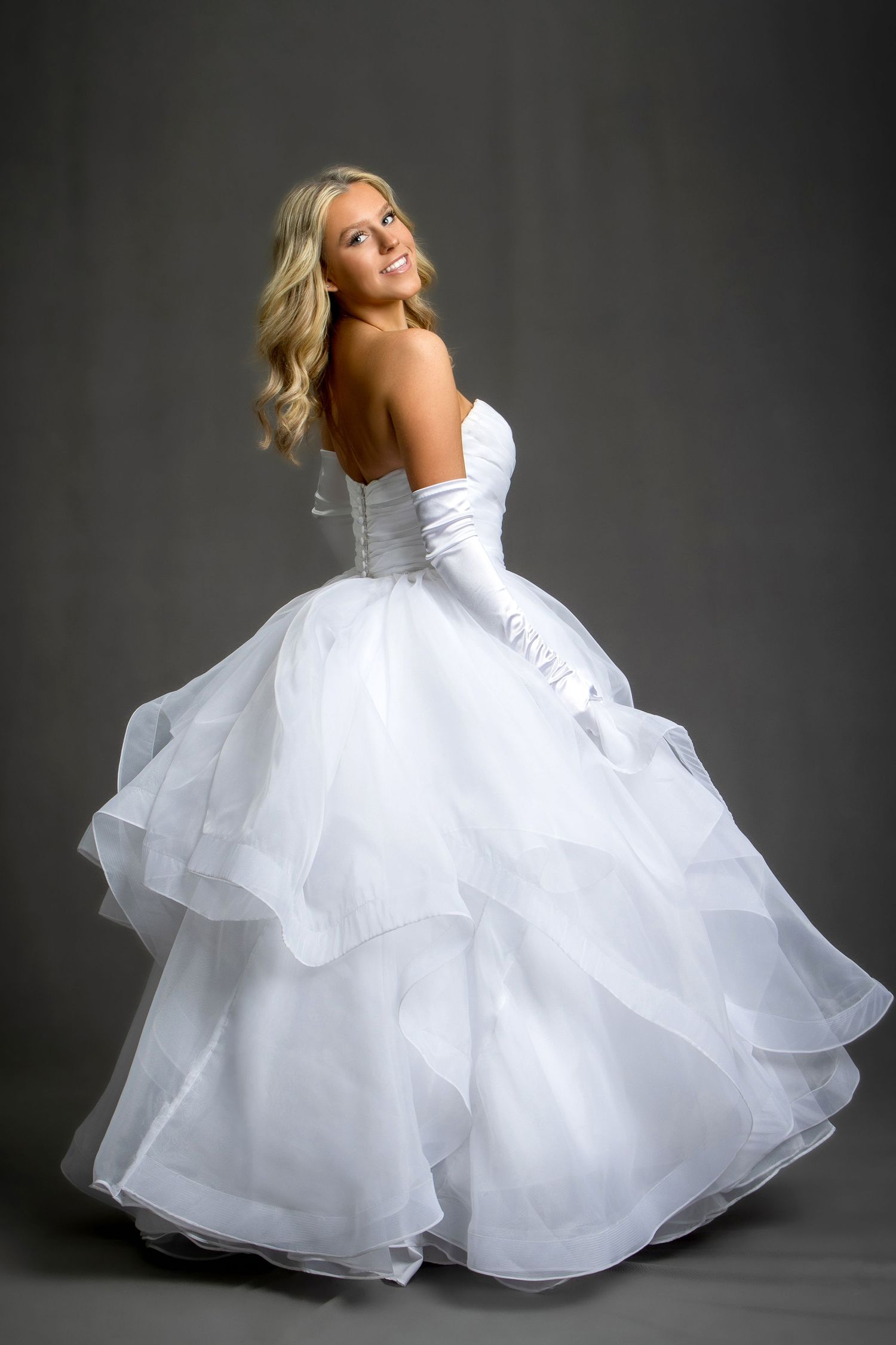 Blonde bride in a strapless white ball gown with layered organza skirt and white gloves, posing gracefully.
