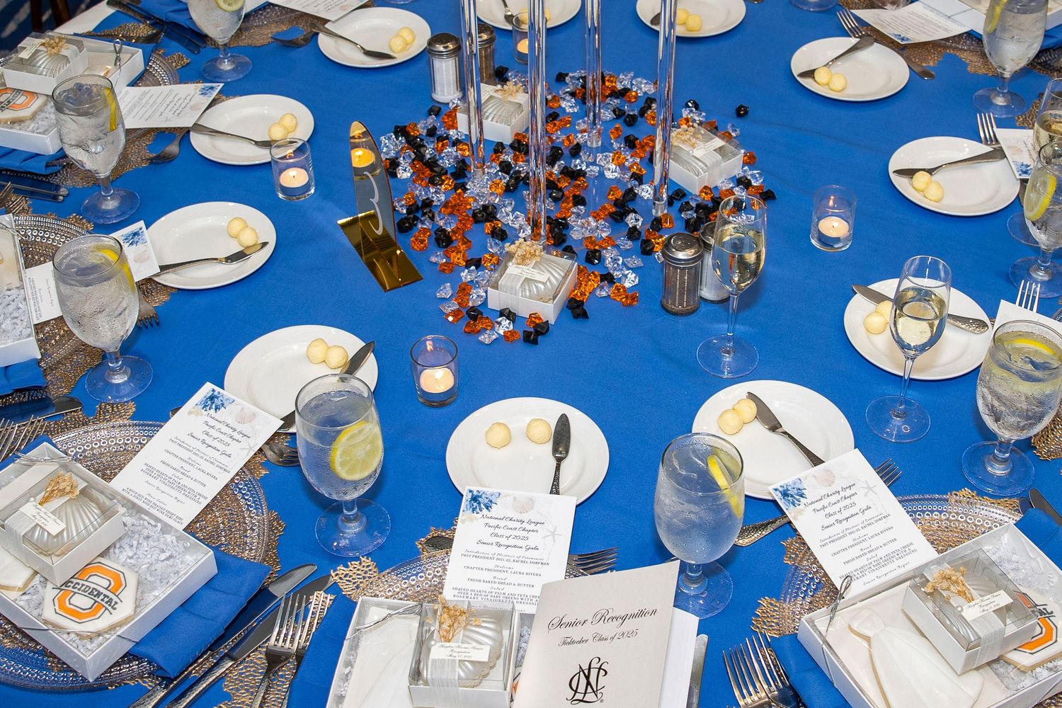 Elegant banquet table with blue tablecloth, white plates, gold cutlery, and colorful candy centerpiece for a formal event.