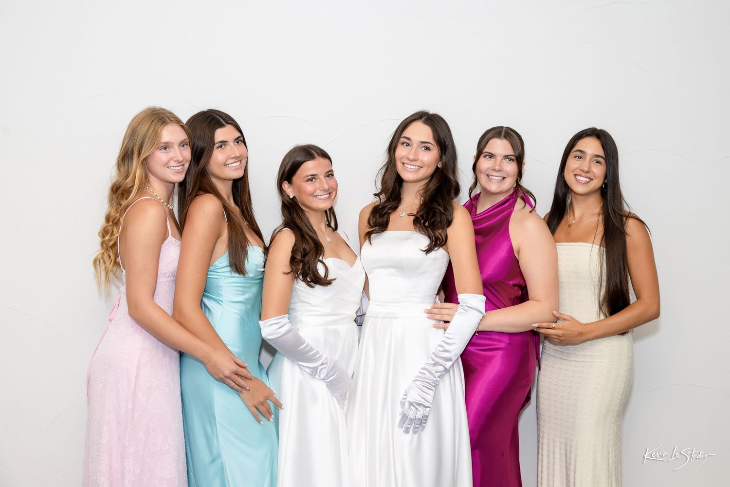 Six young women in elegant formal gowns pose together smiling at a prom or formal event.