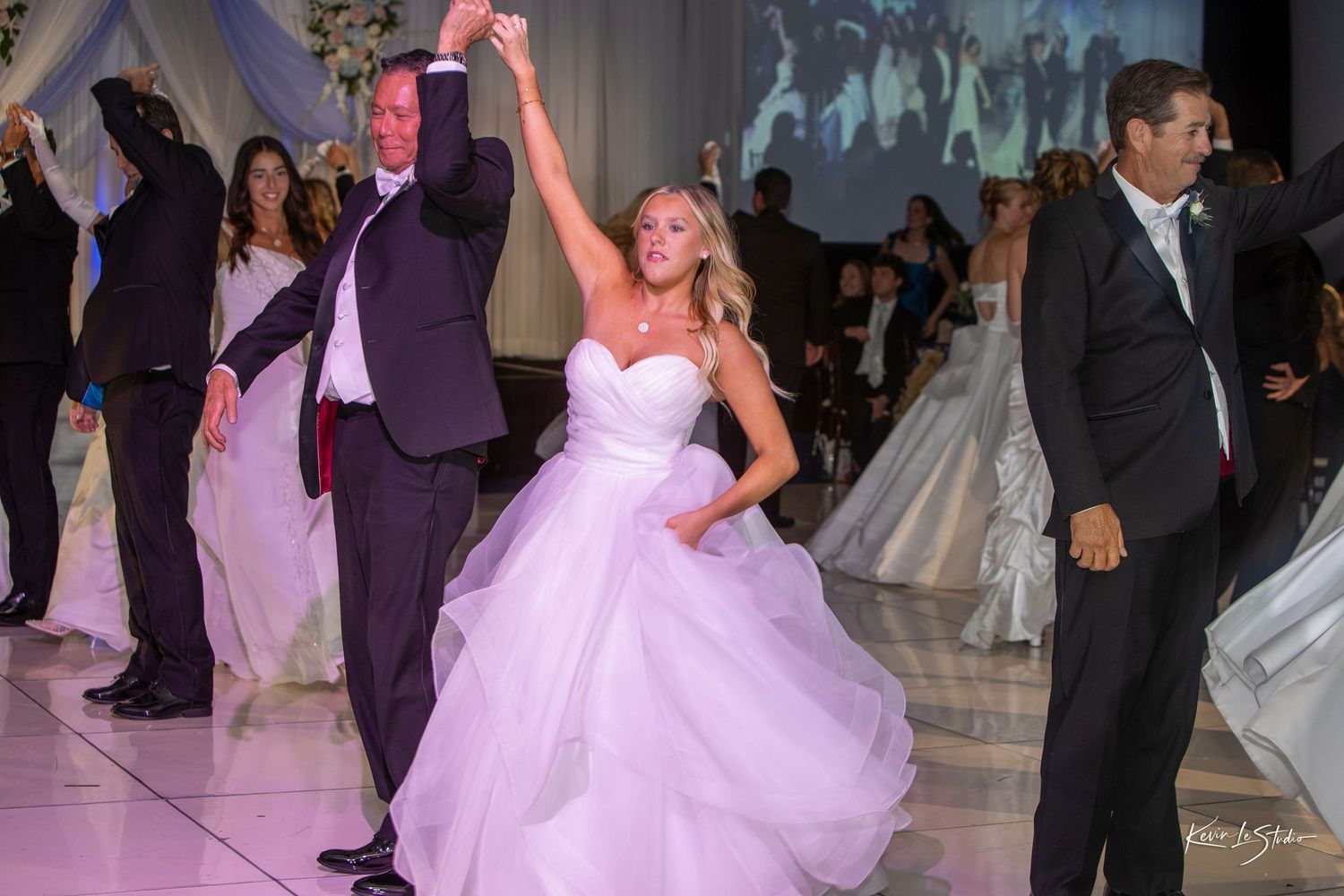 Bride in white gown dancing joyfully with guests at elegant wedding reception on white dance floor.