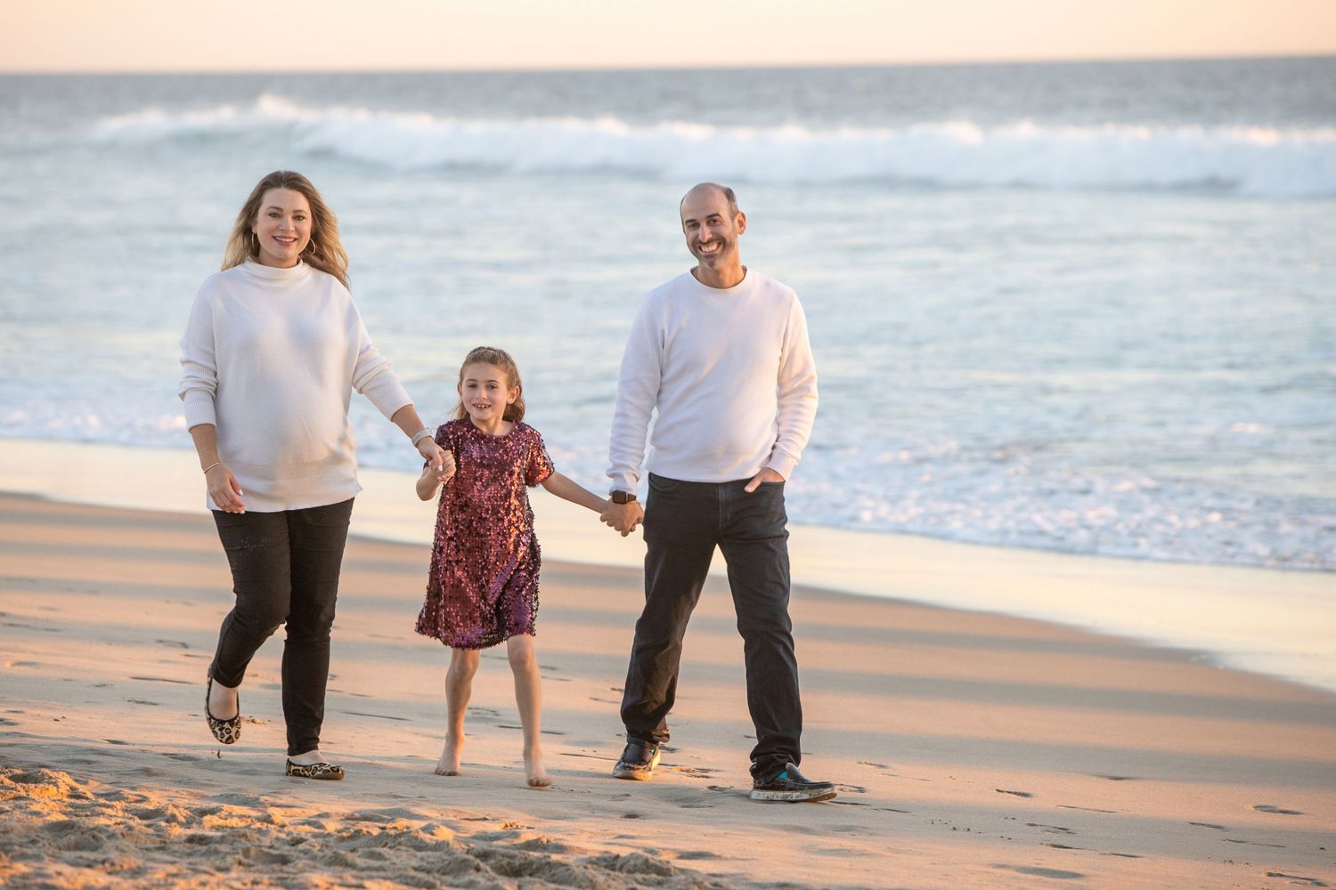 Family of three walking hand-in-hand on a sandy beach at sunset, wearing casual white and colorful outfits.