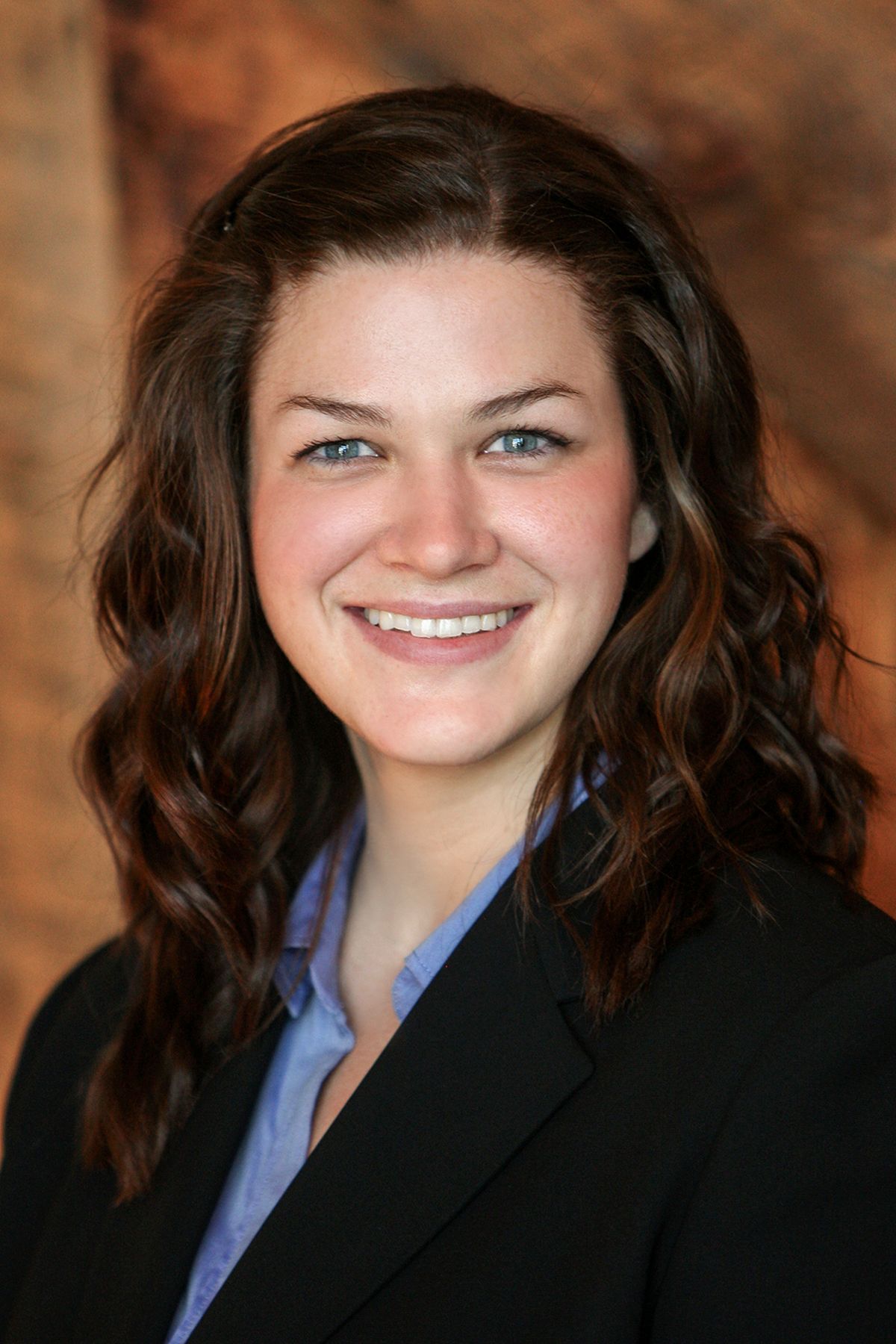 Professional headshot of a young woman with curly brown hair and blue eyes wearing a black blazer.
