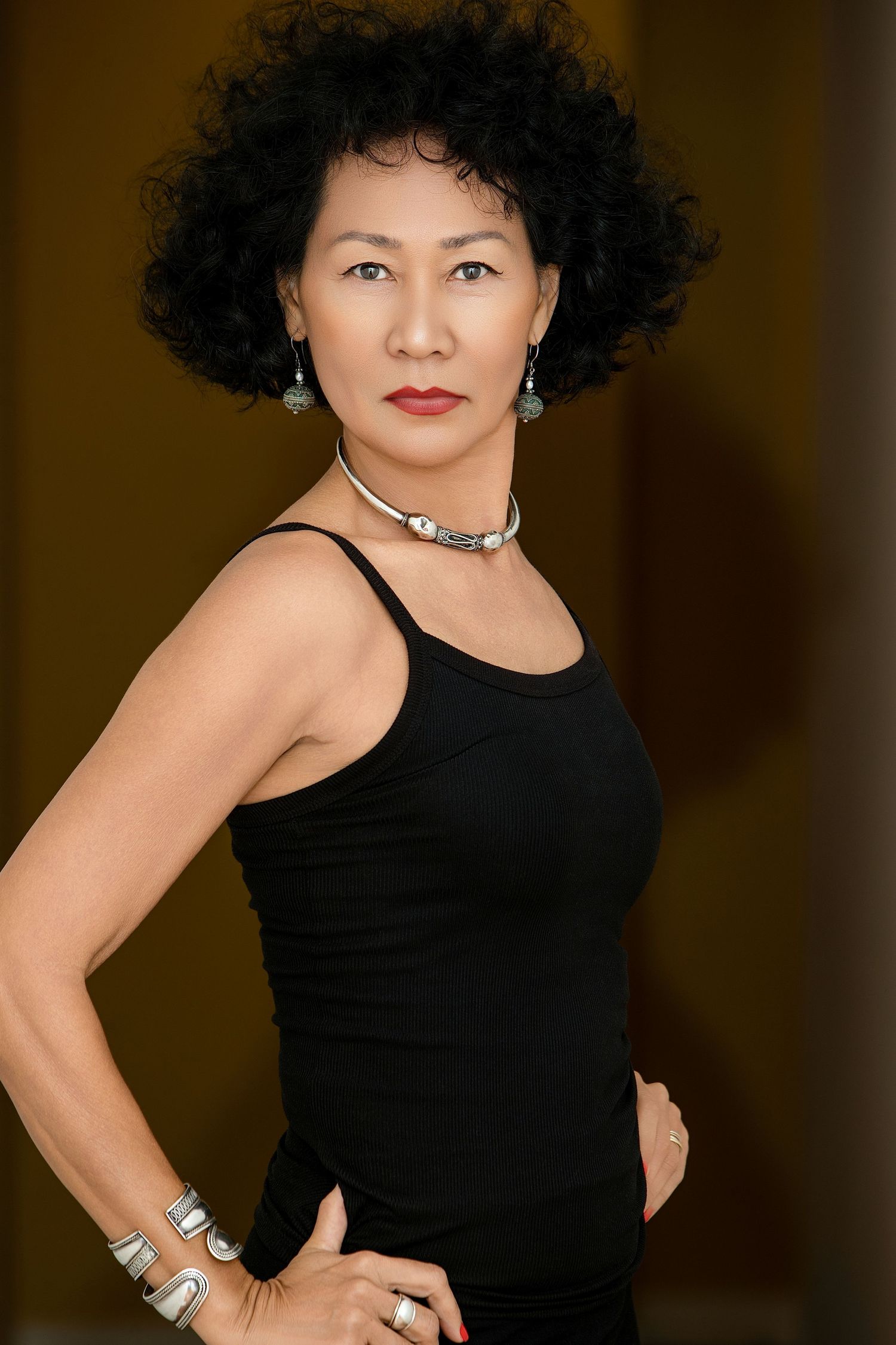 Confident mature Asian woman in black tank top with silver jewelry, hand on hip, studio portrait.
