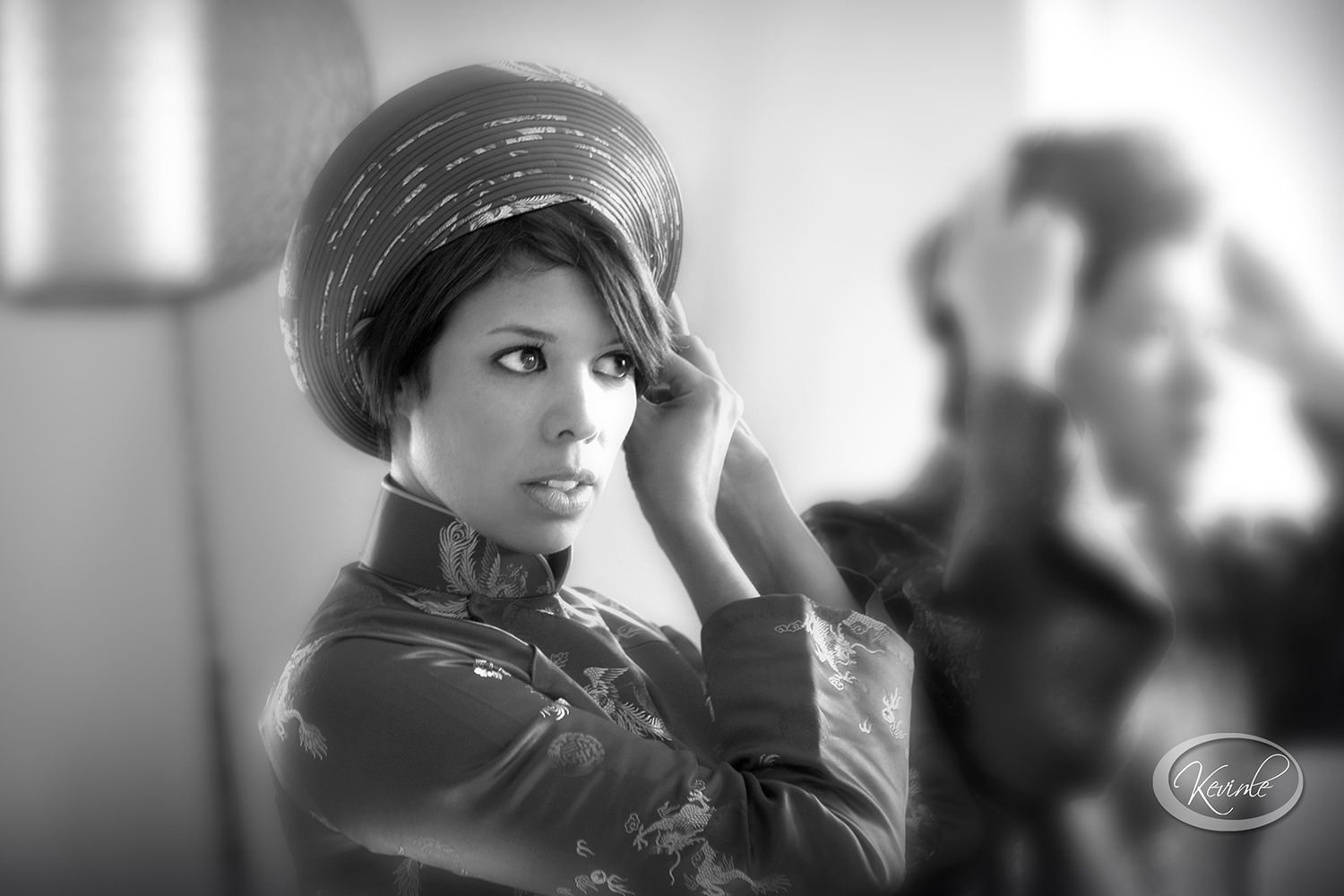 Black and white portrait of woman in traditional Vietnamese ao dai adjusting hat, looking back.