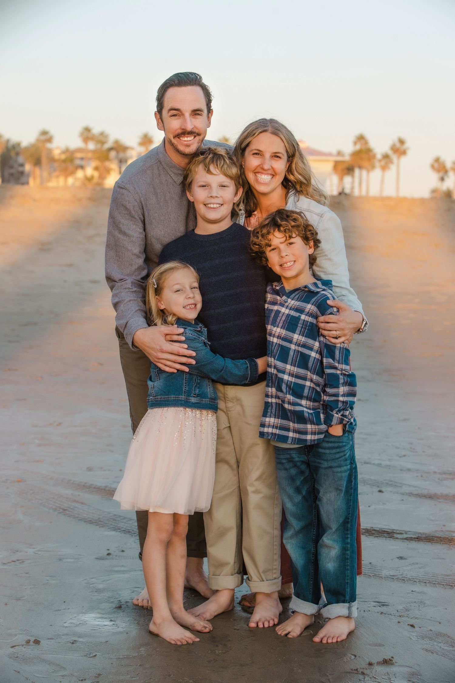 Happy family of five poses together barefoot on a sandy beach during golden hour photography session.